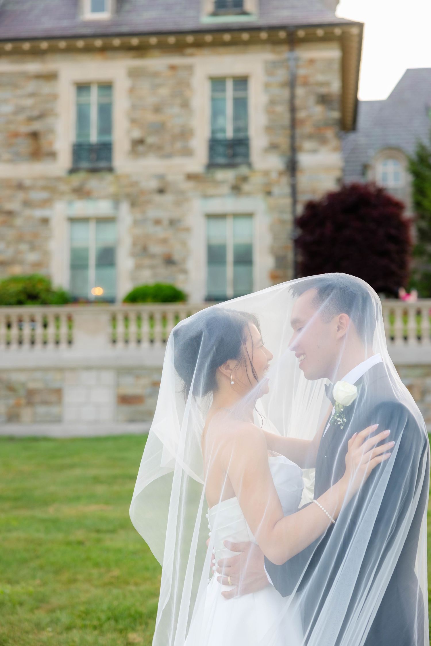A bride and groom share a romantic moment under a flowing veil outside a stone mansion.