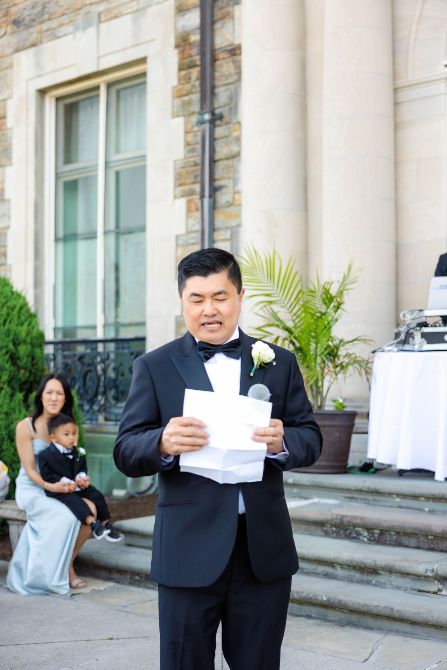 Groom in black tuxedo reading vows from paper during outdoor wedding ceremony at stone estate.