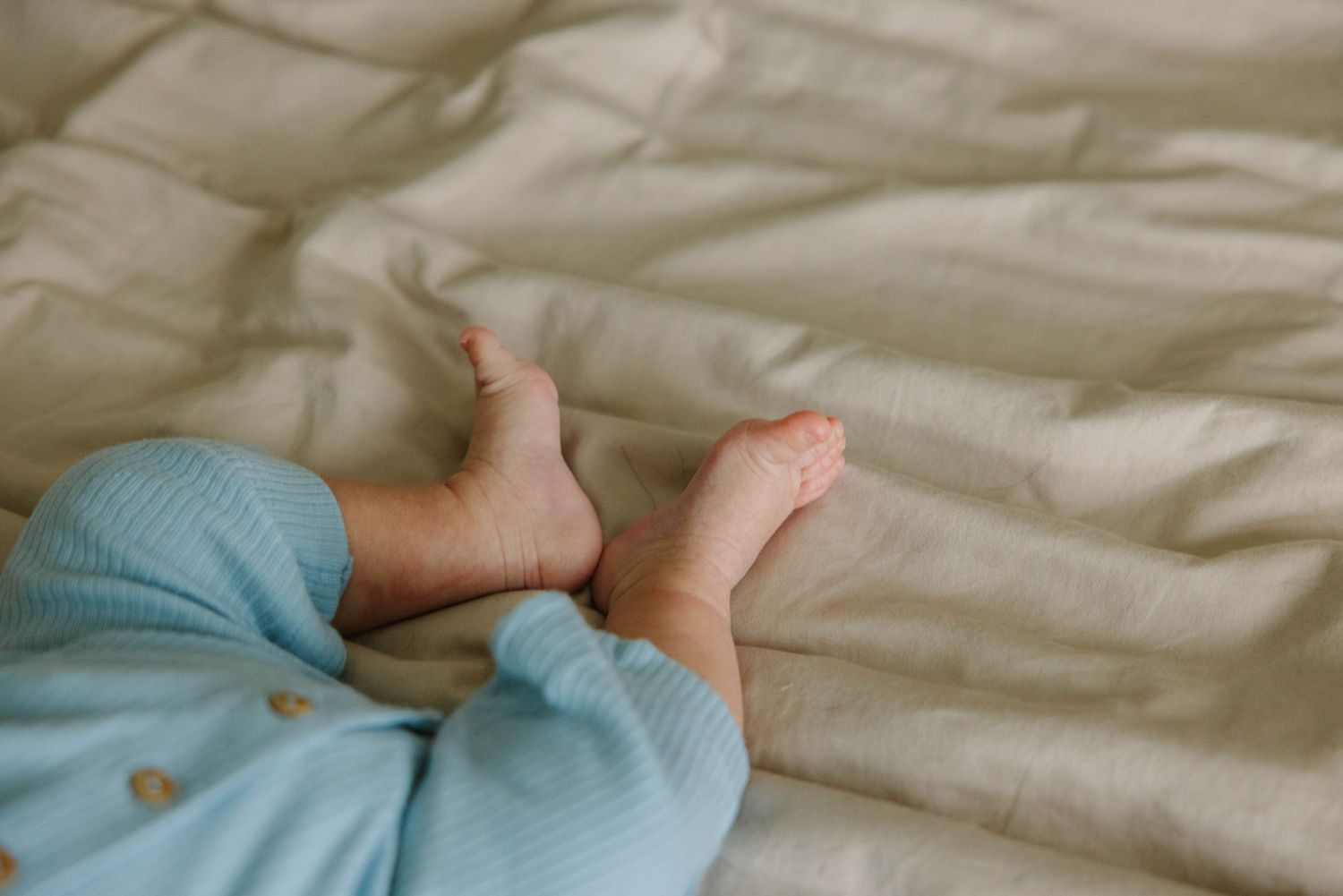 Close-up of baby feet in light blue pajamas resting on a soft white linen bed.