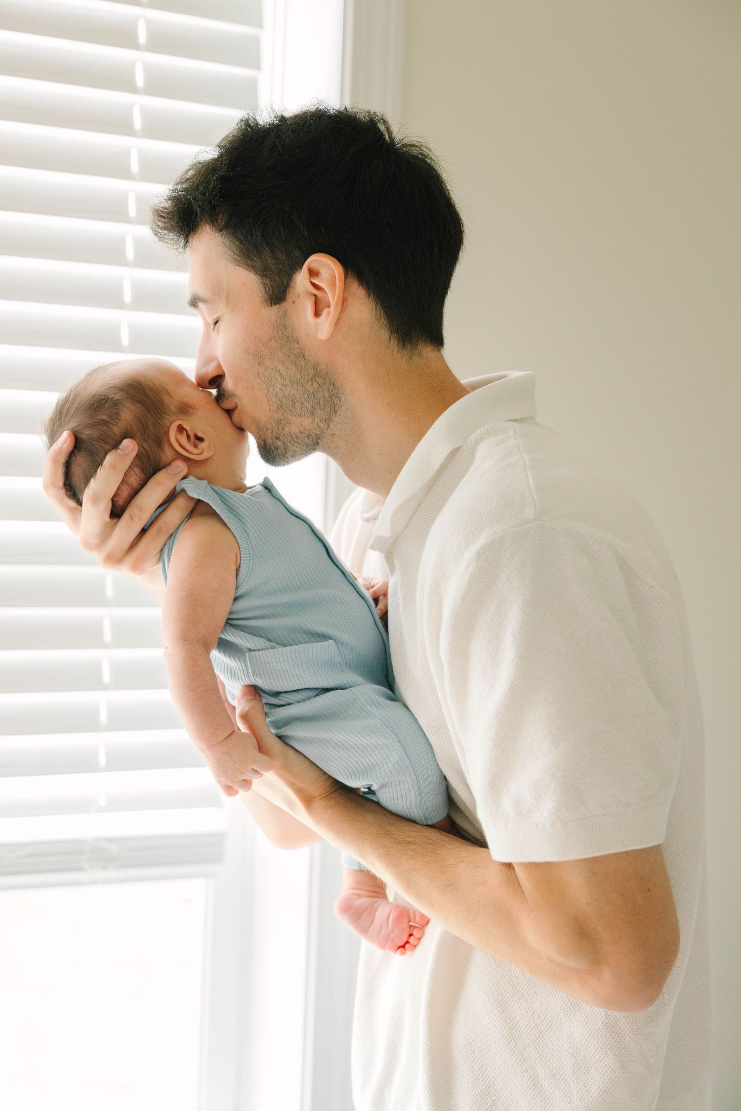 A loving father gently kisses his newborn baby dressed in blue, standing by a bright window with white blinds.