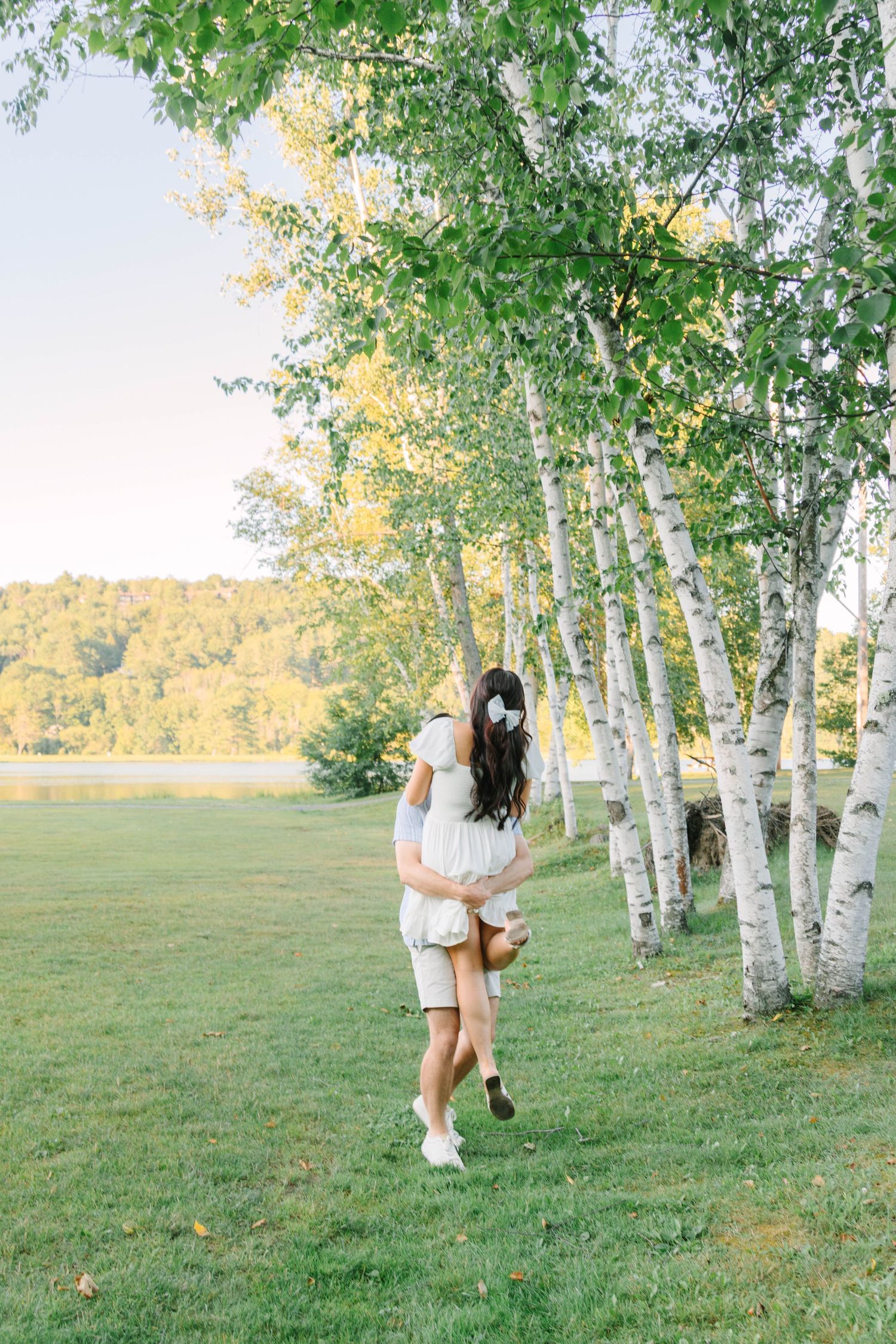 Couple embracing near birch trees on green grass by a lake, woman in white dress lifted by man in summer outdoor setting.