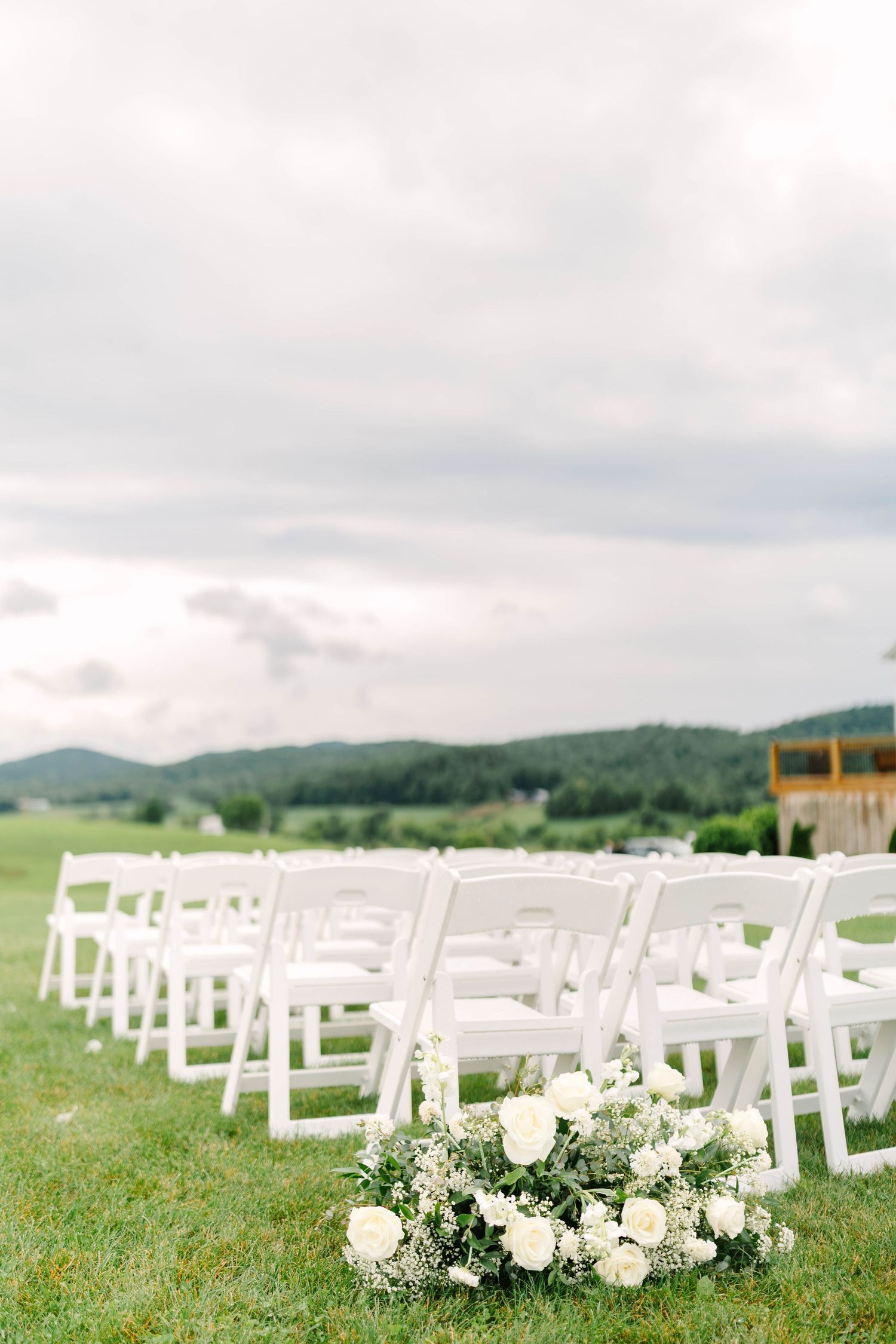 White folding chairs and floral arrangements set up on a green field for an outdoor wedding ceremony.