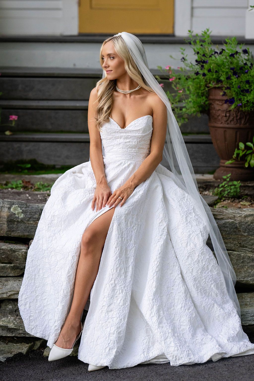 Bride in strapless lace ball gown with veil sitting on stone steps outdoors.