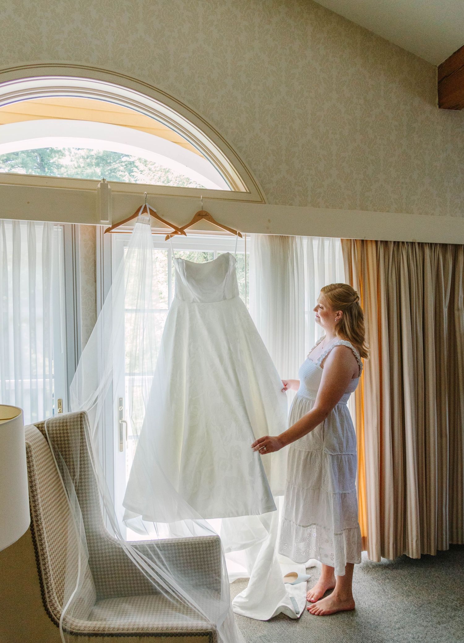Blonde bride admires her strapless white wedding gown hanging by an arched window in a hotel room.