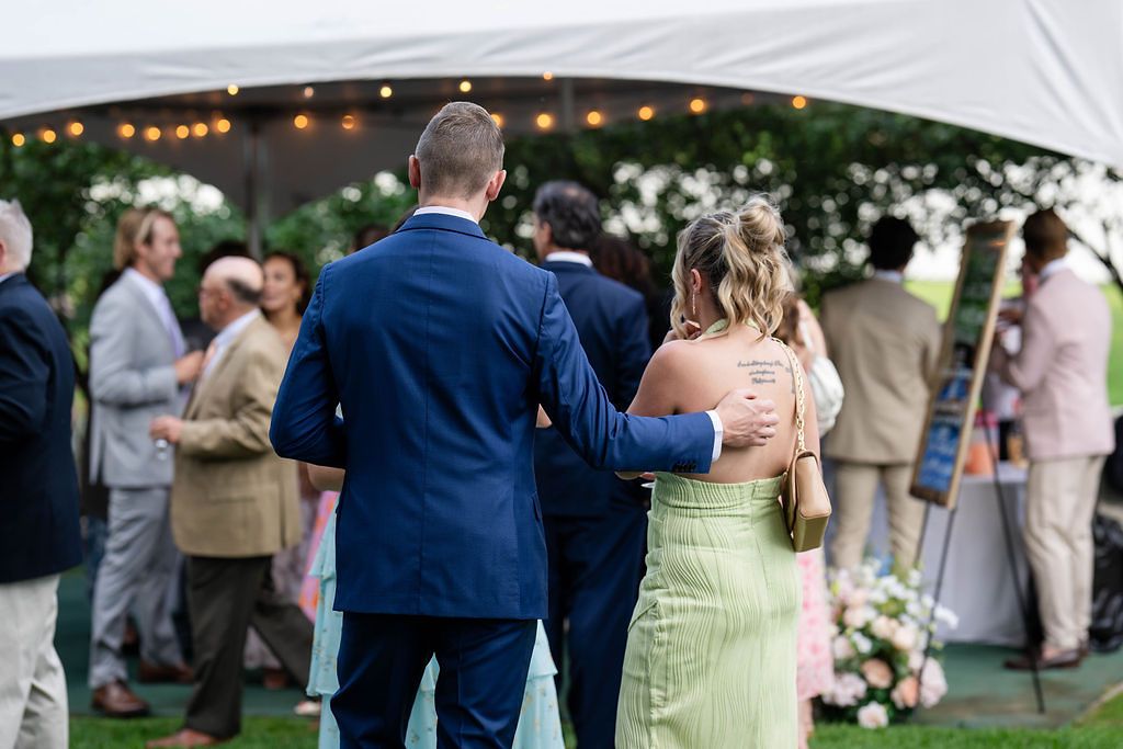 Guests in formal attire walk toward a decorated outdoor wedding tent with string lights and floral arrangements.