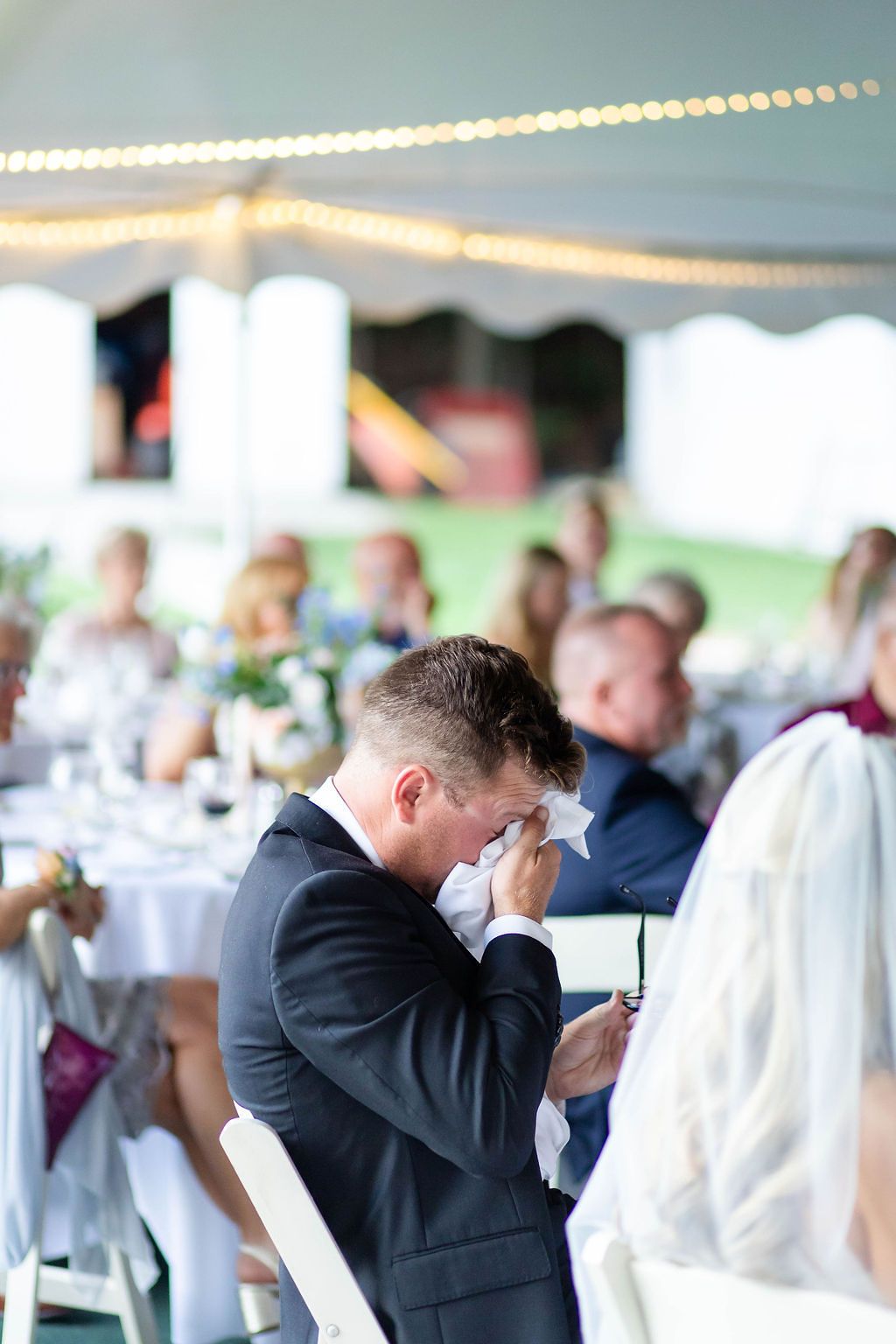 Emotional groom wipes tears during wedding ceremony under white tent with guests seated.