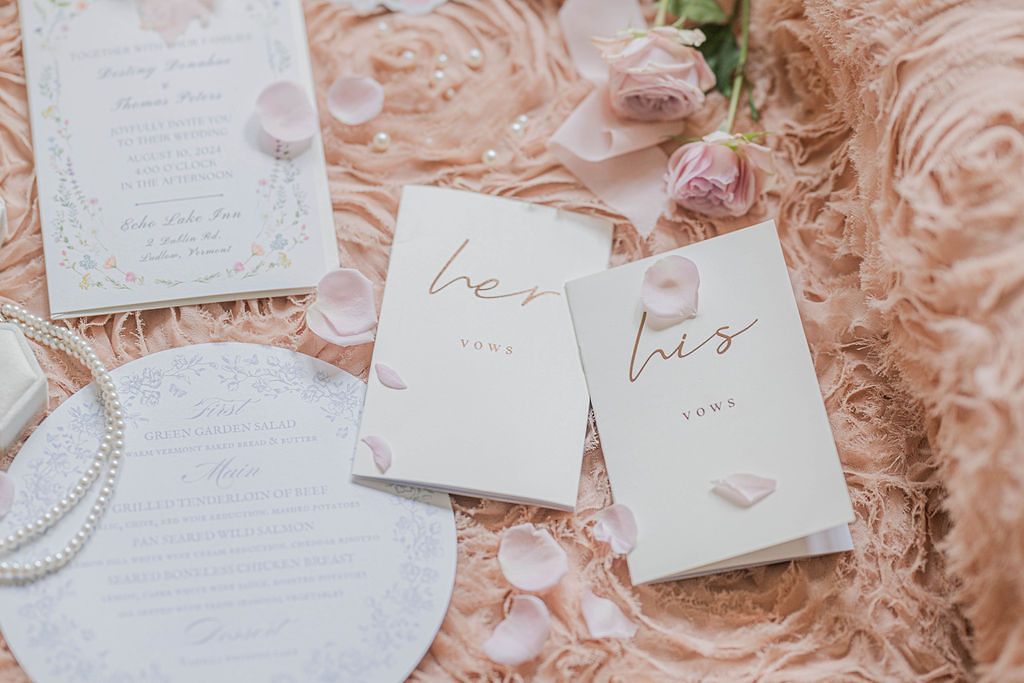 Elegant wedding stationery flatlay with his and hers vow books, floral invitation, pearl bracelet, and pink roses on blush fabric.