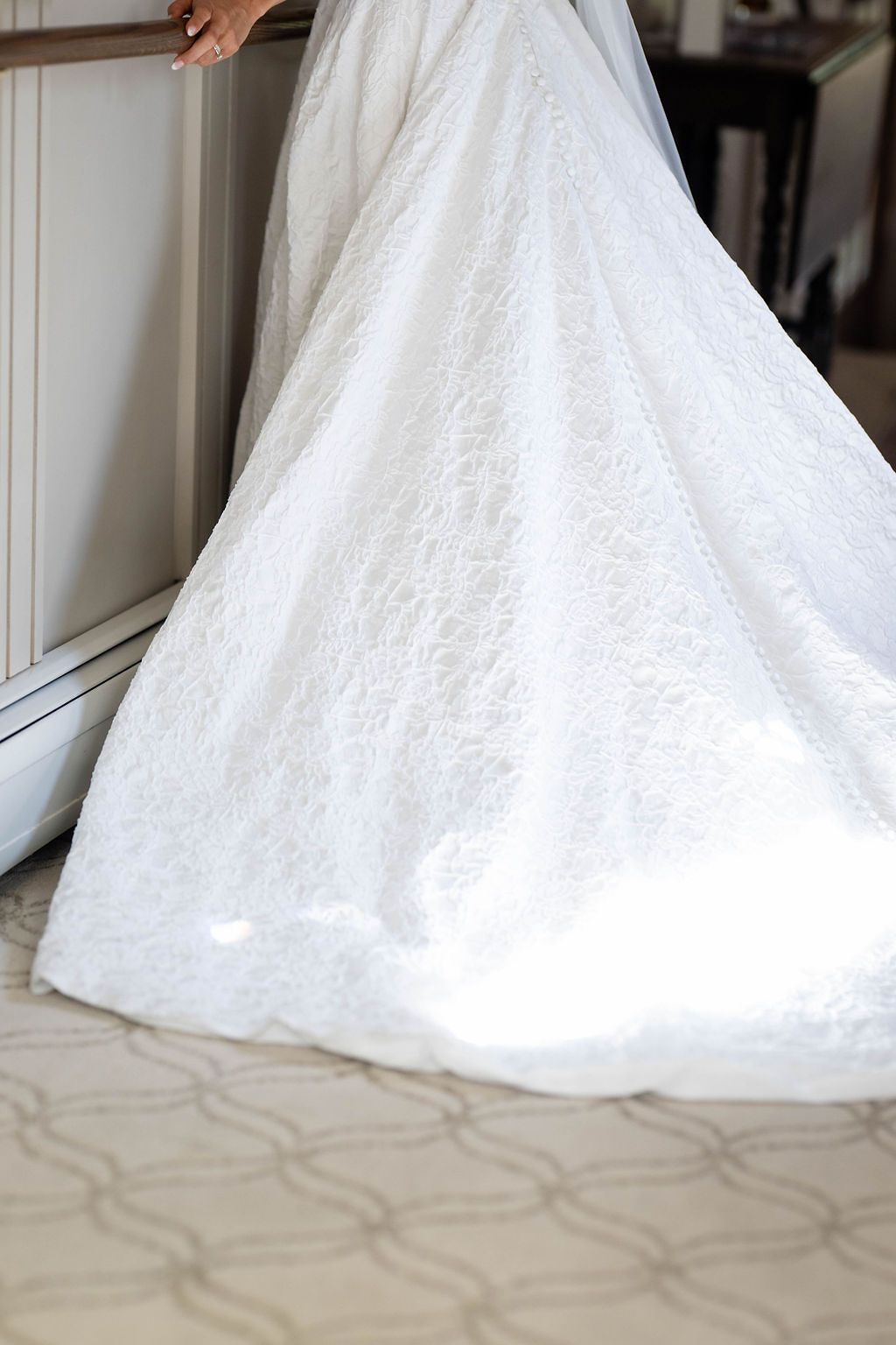 Close-up of a bride's lace wedding gown train spreading elegantly across a carpeted floor near a window.