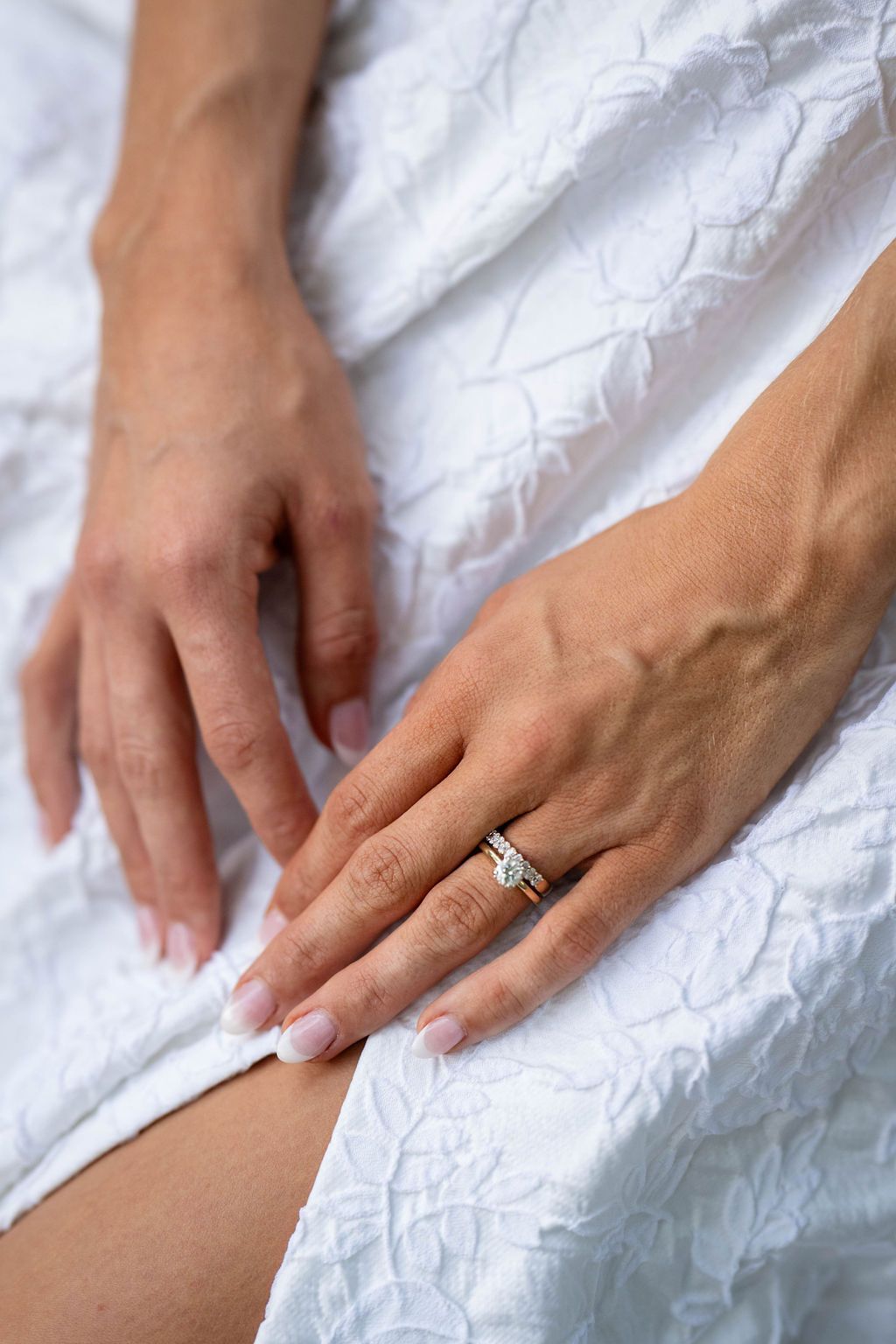 Close-up of bride's hands resting on white lace wedding gown showing gold diamond engagement ring.