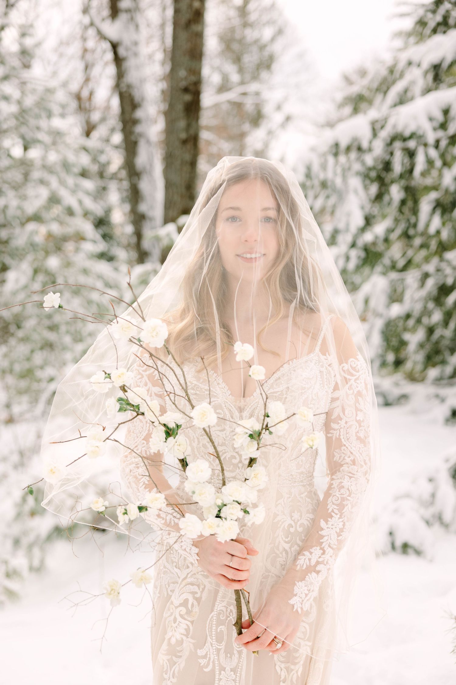 Bride in lace wedding dress with veil holding white flowers in winter snow.