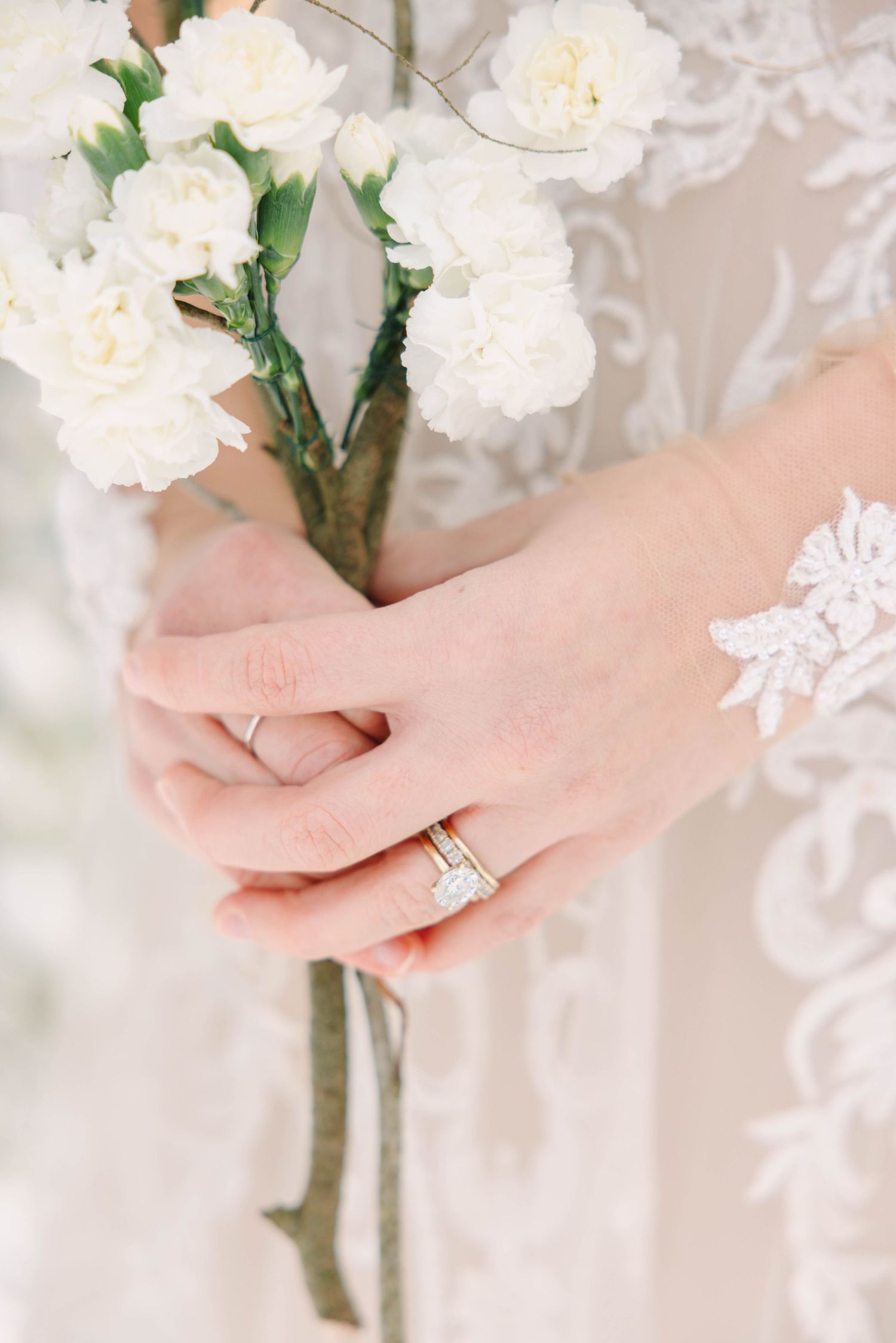 Close-up of bride's hands holding white carnation bouquet with diamond engagement ring.