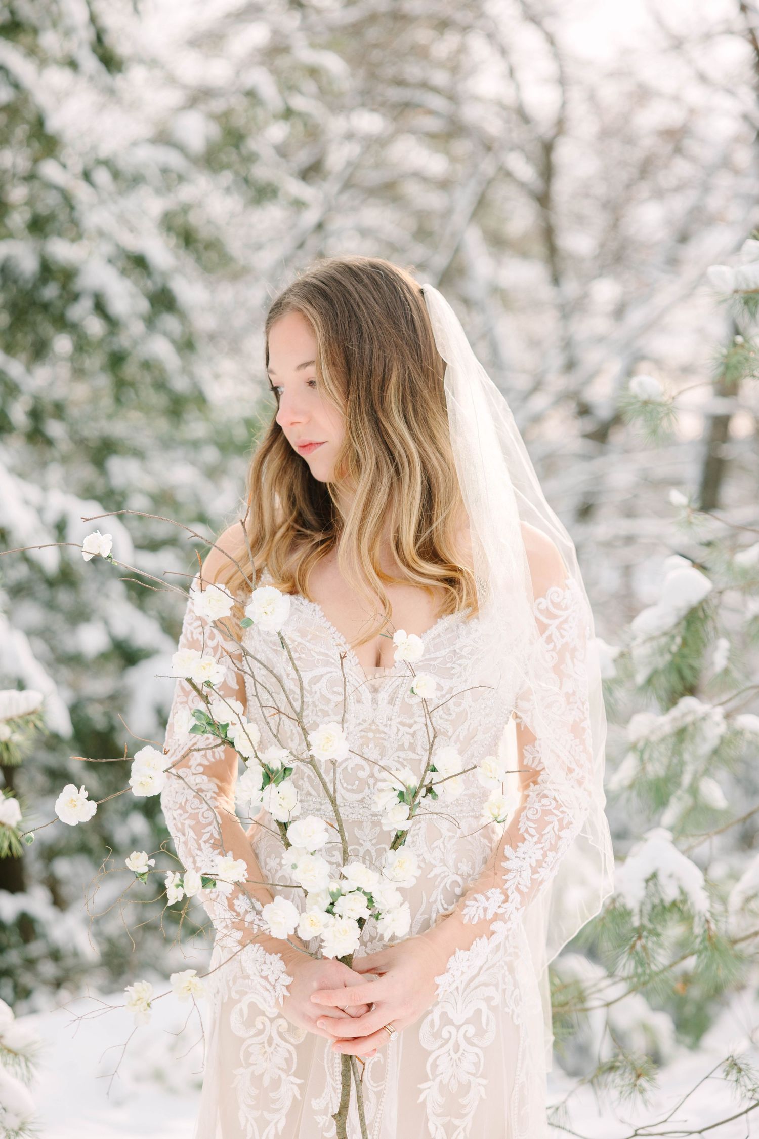 Bride in lace gown and veil holds white flowers in snowy winter forest setting.