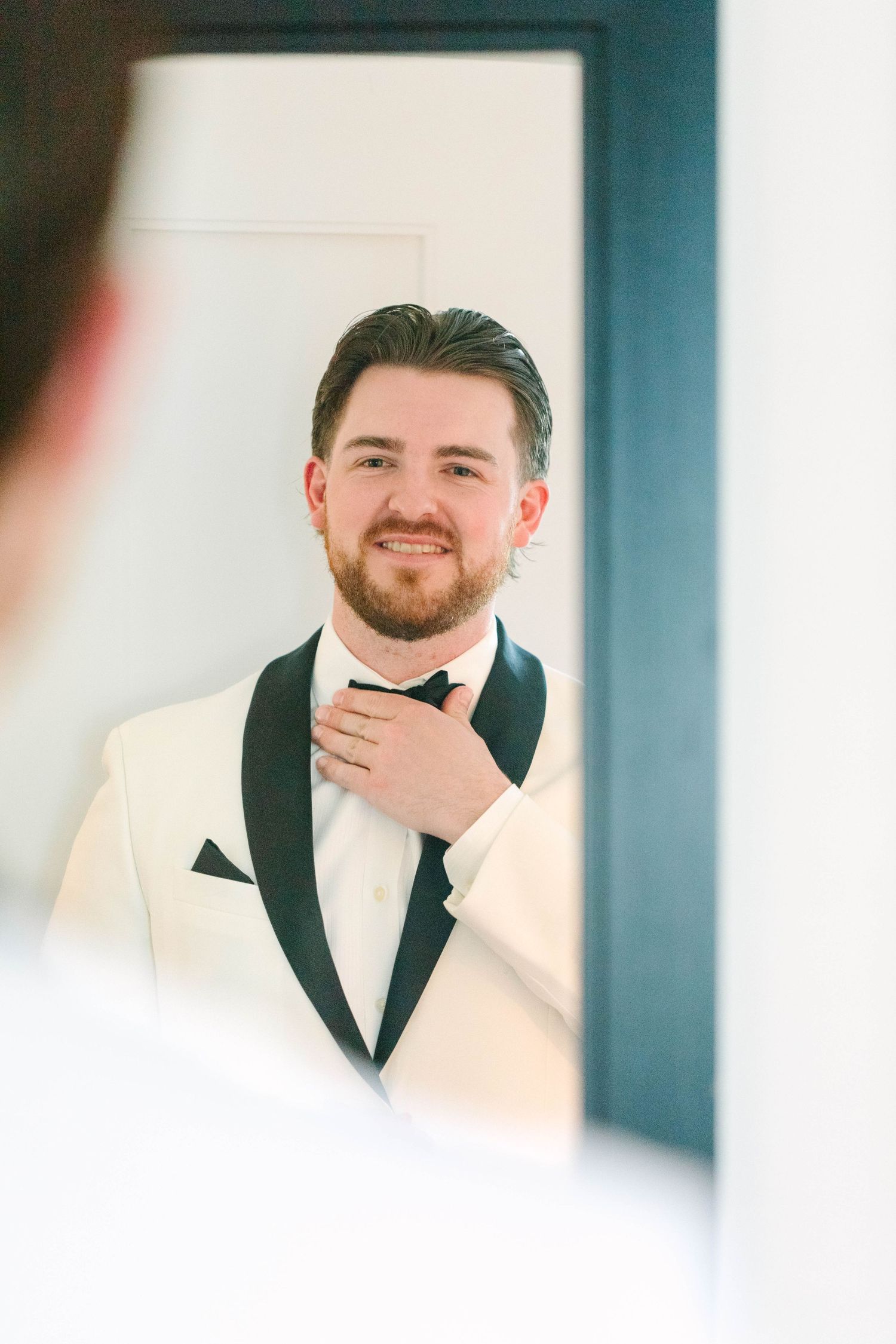 Groom in white tuxedo jacket adjusting black bow tie while looking in mirror before wedding.