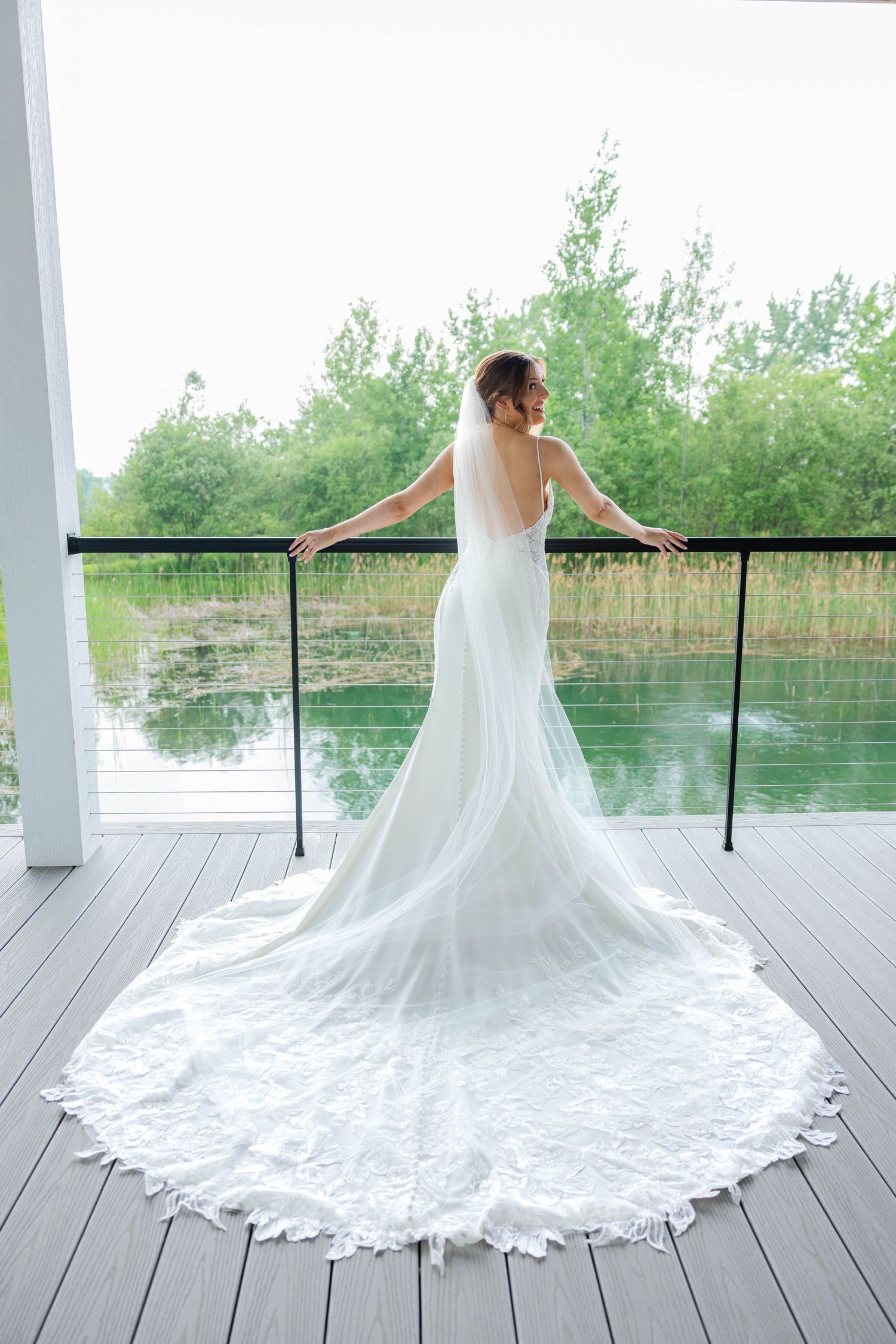 Bride in backless lace wedding gown with long train stands on balcony overlooking serene pond.