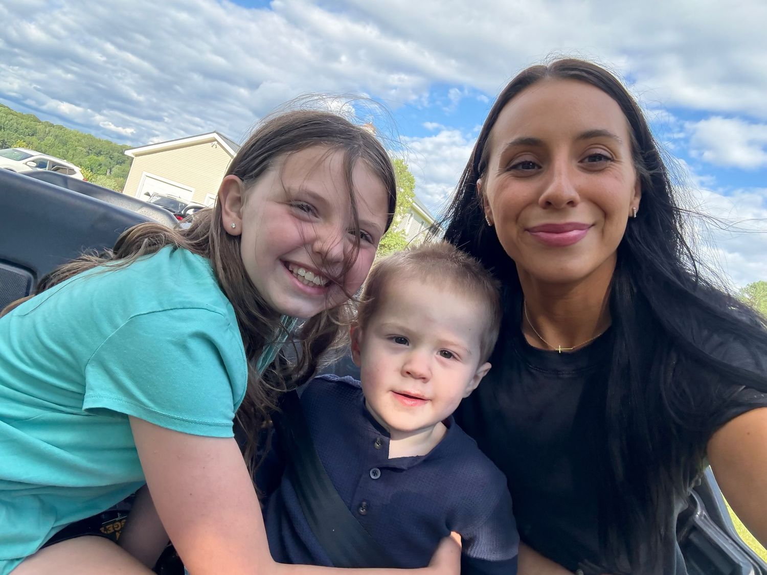 A woman and two children smile together in a selfie outdoors under a partly cloudy blue sky.