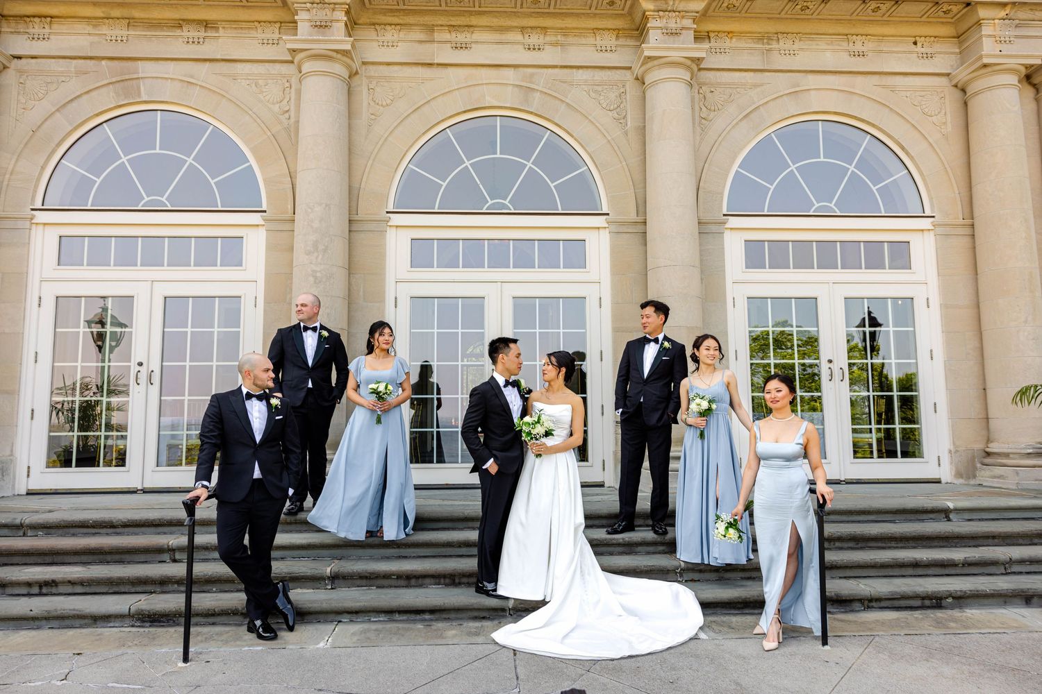 Wedding party poses on elegant mansion steps, bride in white gown with groomsmen in tuxedos and bridesmaids in blue dresses.