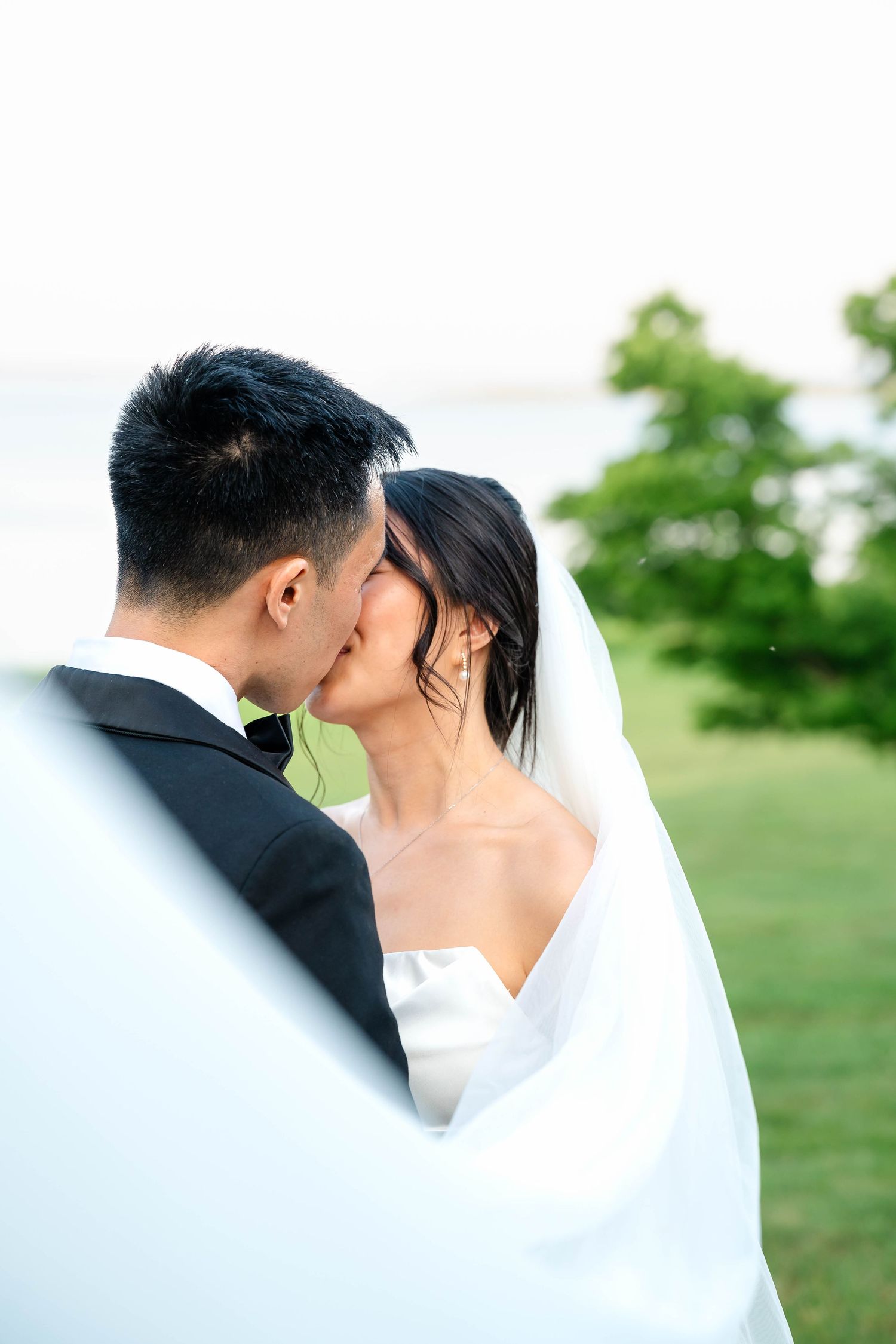 Bride and groom share a tender kiss wrapped in flowing white veil with green trees and water behind.