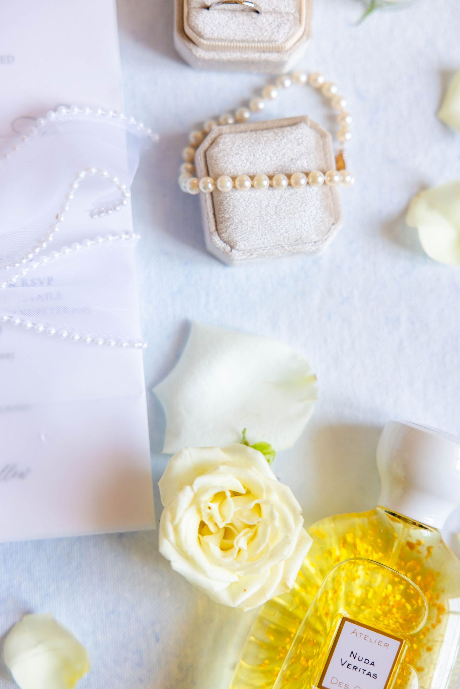 Elegant bridal flat lay with pearl jewelry, ring box, white roses, and Nuda Veritas perfume on soft blue background.