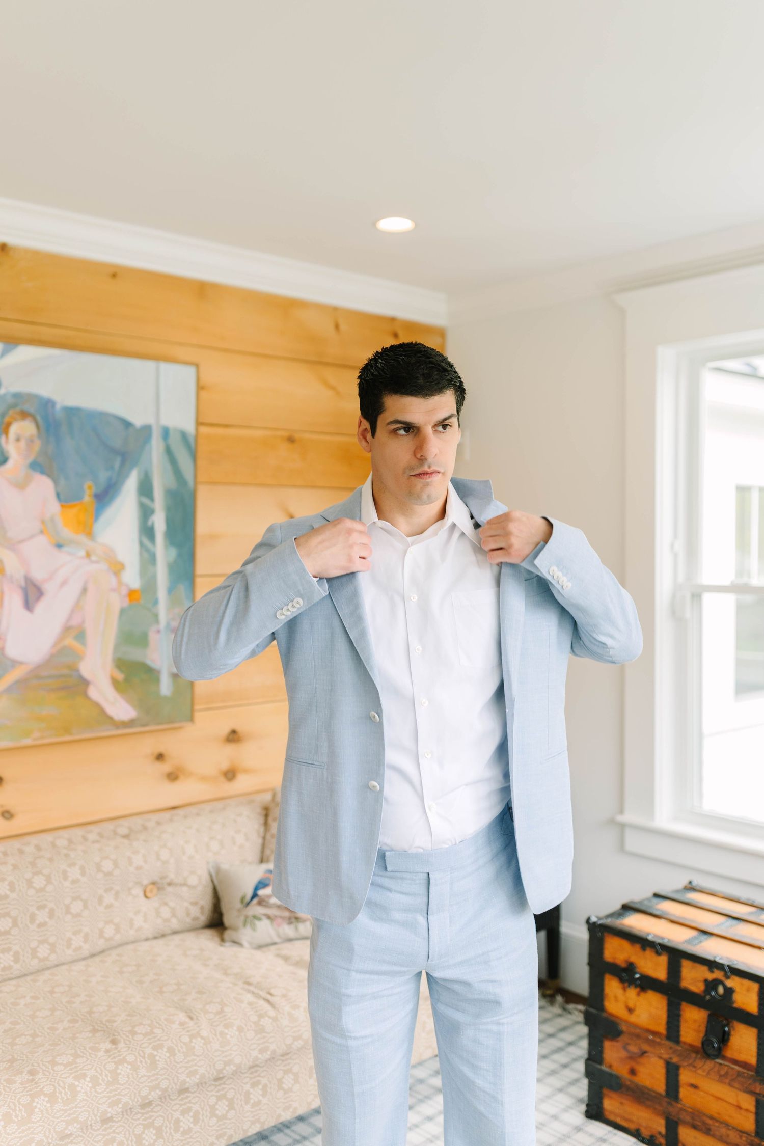Groom adjusts his light blue suit jacket while standing in a well-lit bedroom before his wedding.