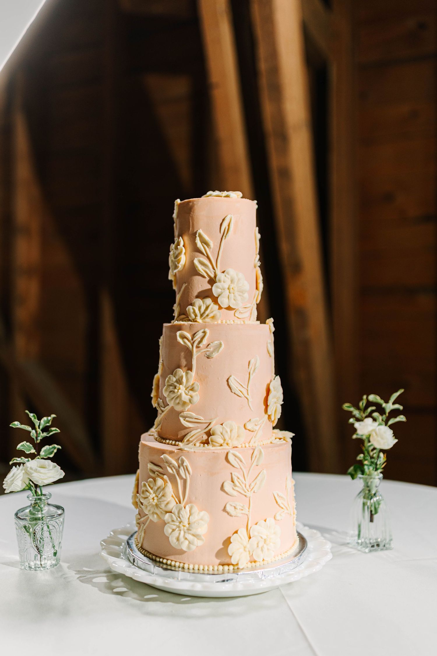 Elegant three-tier blush pink wedding cake with gold floral details on white table in barn.