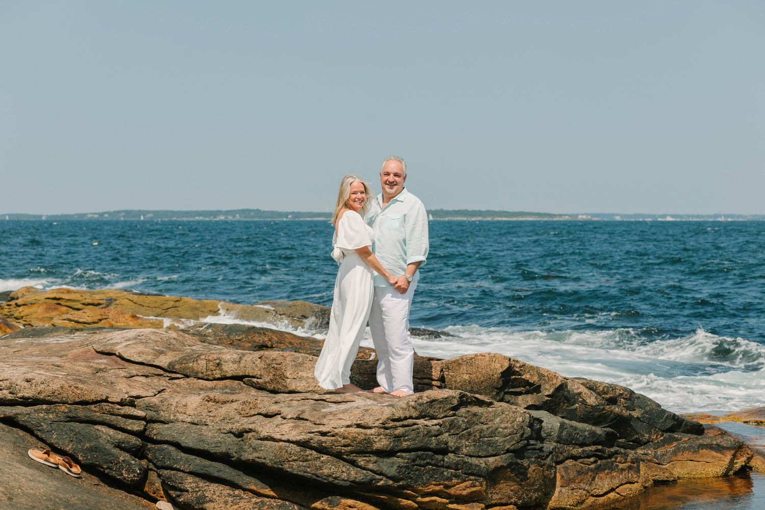 Elderly couple in white outfits embracing on rocky New England coastline with blue ocean waves behind them.