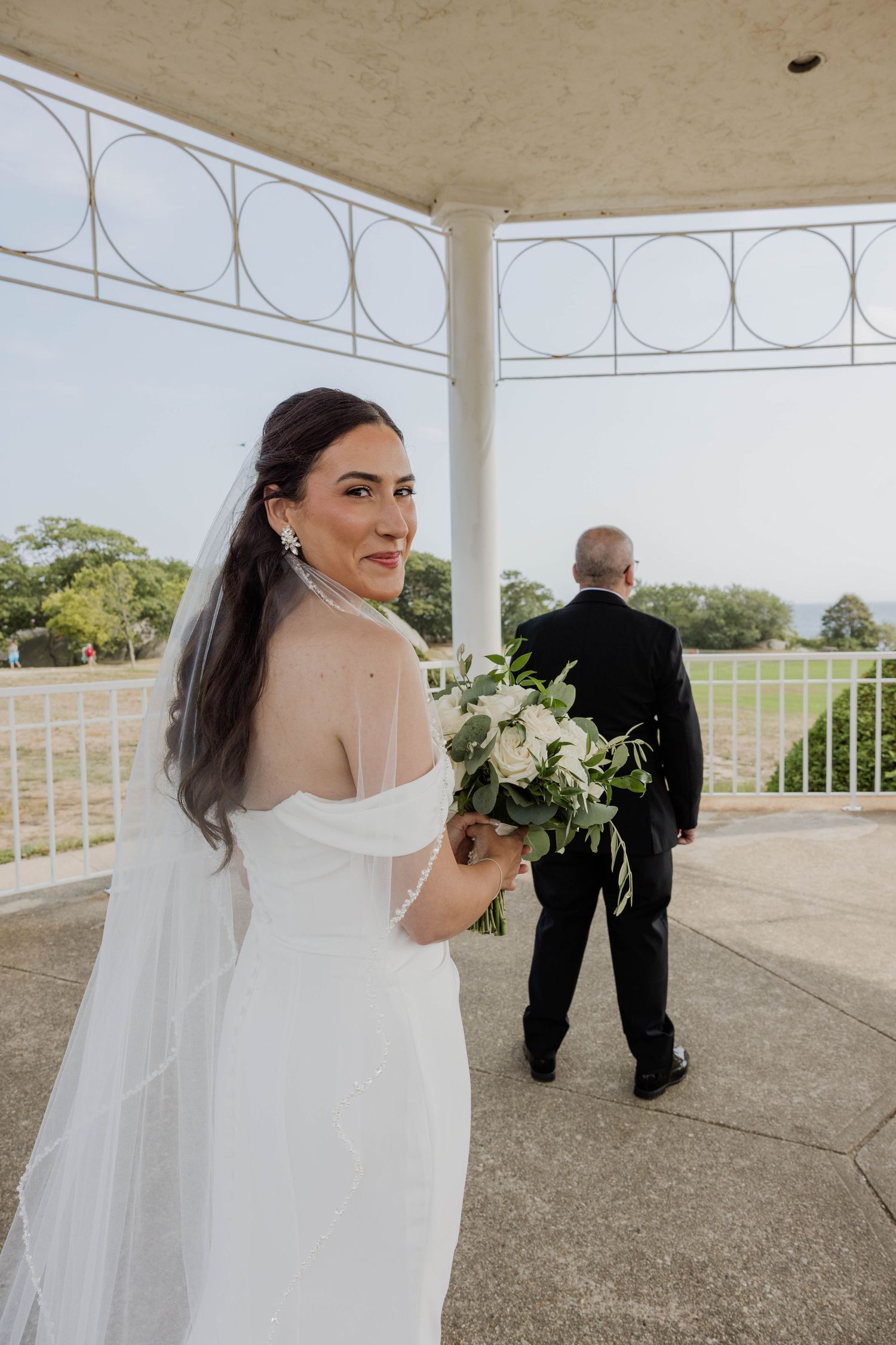 Bride in off-shoulder white gown holding bouquet looks back while groom stands at outdoor gazebo.