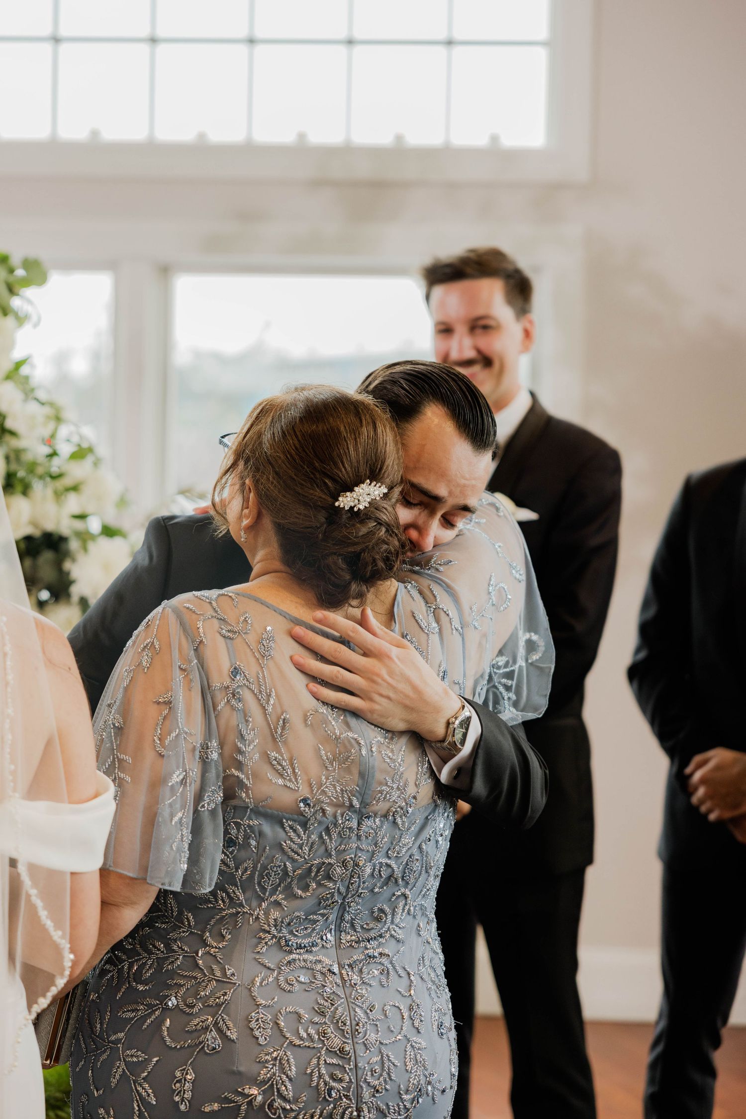 Groom embracing a woman in a blue lace dress at a wedding ceremony while a smiling guest looks on.