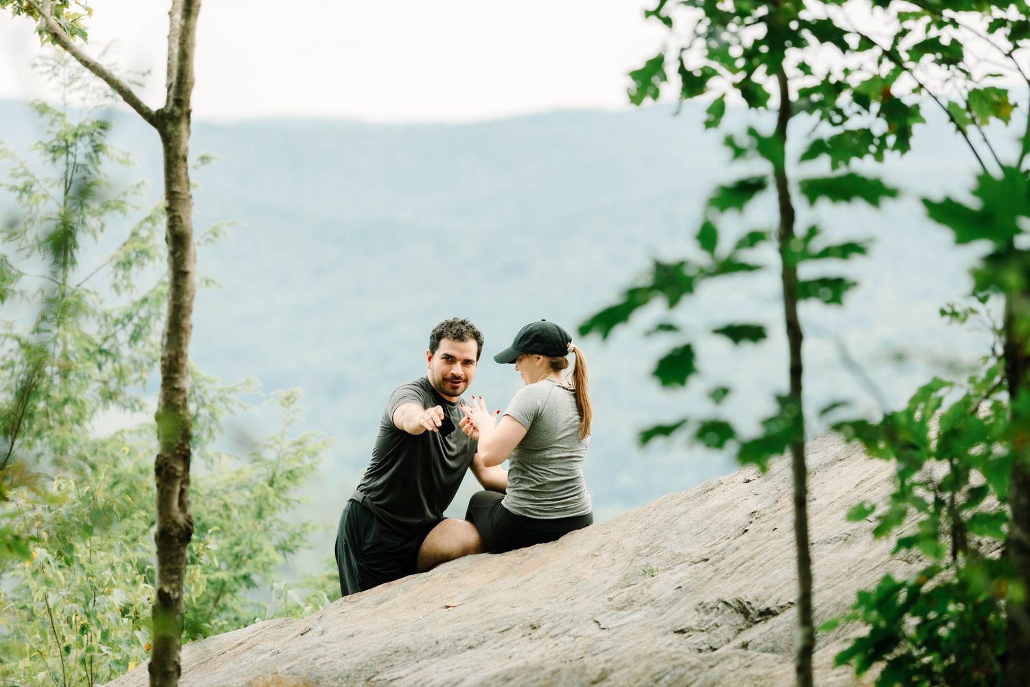 Couple sitting on a rocky ledge surrounded by lush trees with a scenic mountain valley view in the background.