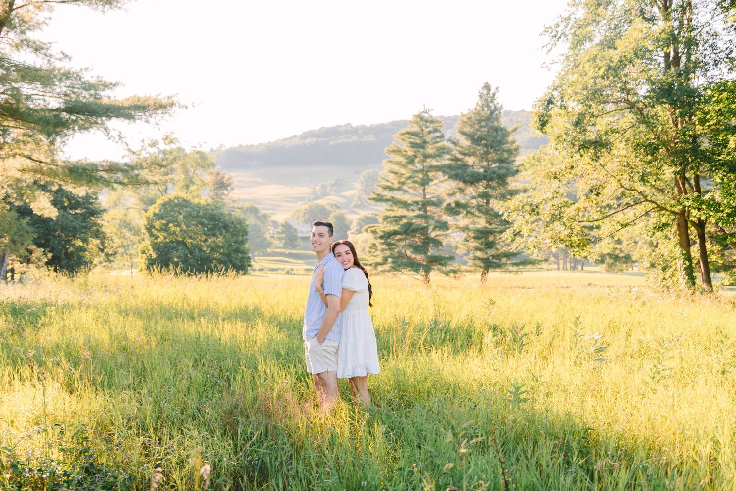 Couple embracing in a sunlit meadow surrounded by lush green trees and rolling hills during golden hour.
