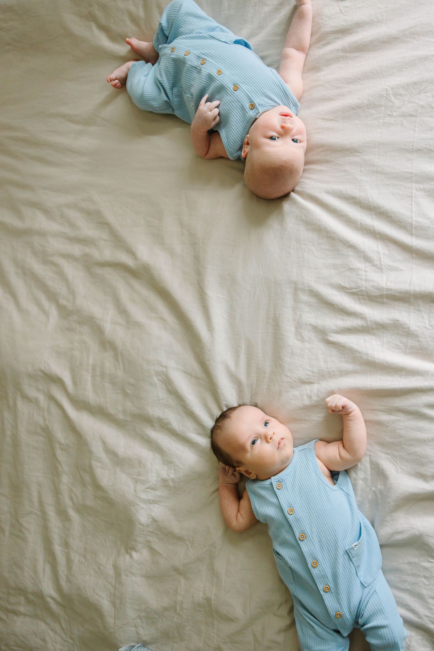 Two newborn twin babies in matching blue rompers lying on a beige linen bed from above.