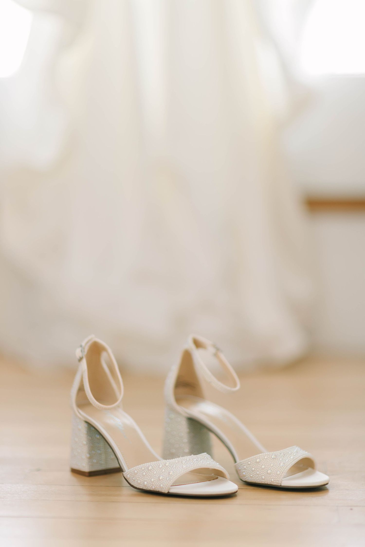 Ivory block heel bridal sandals with ankle strap on wood floor before wedding gown.