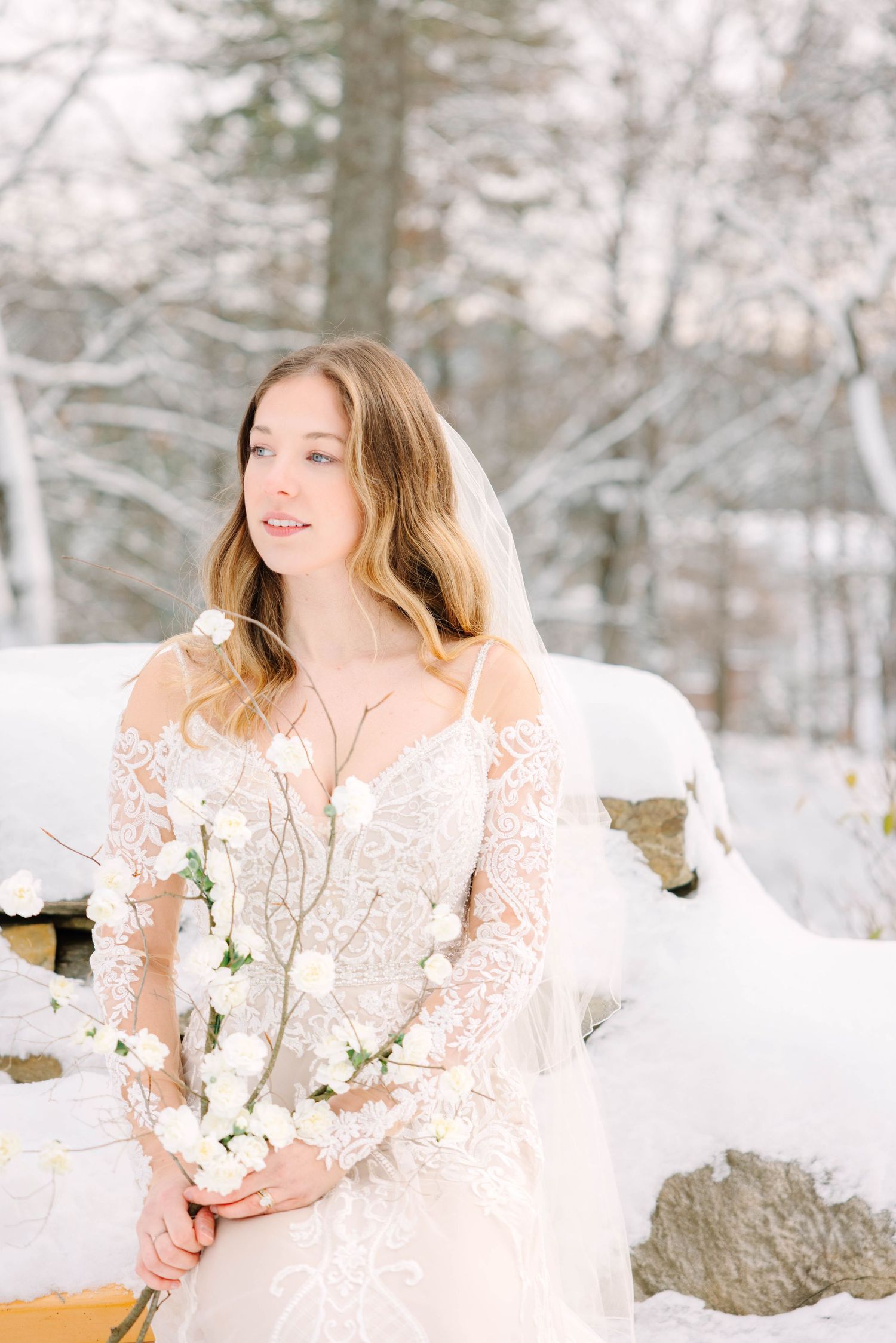 Bride in off-shoulder lace gown and veil holds white floral branch while seated in snowy landscape.