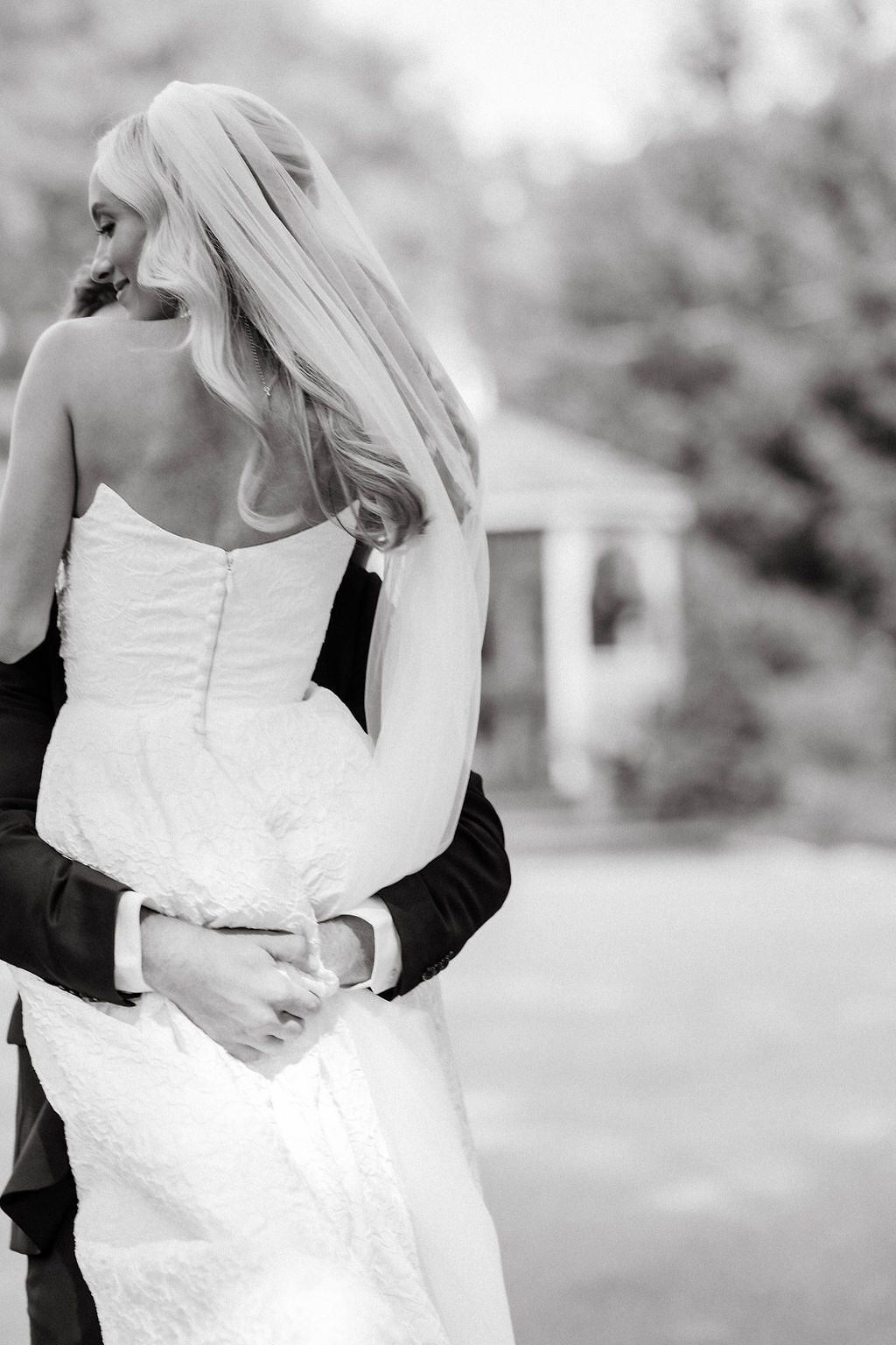 Black and white photo of groom embracing bride in strapless lace wedding gown outdoors.
