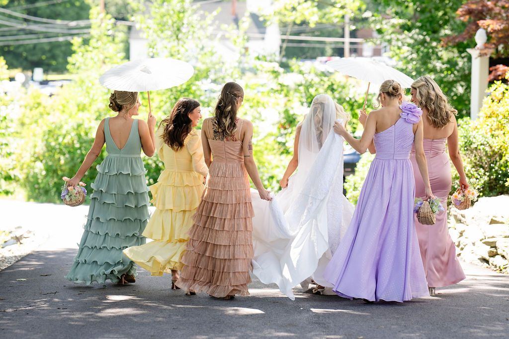 Bride and four bridesmaids in colorful ruffled gowns walk together from behind on outdoor pathway.