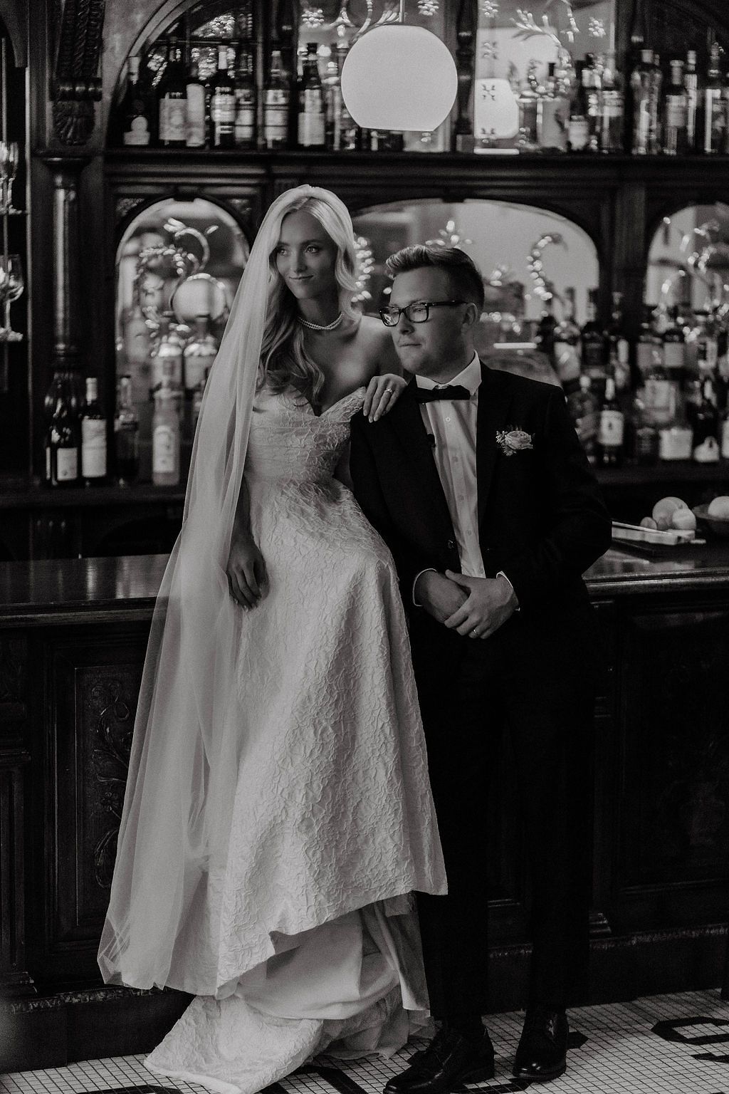 Black and white portrait of bride and groom posing elegantly in front of a classic bar setting.