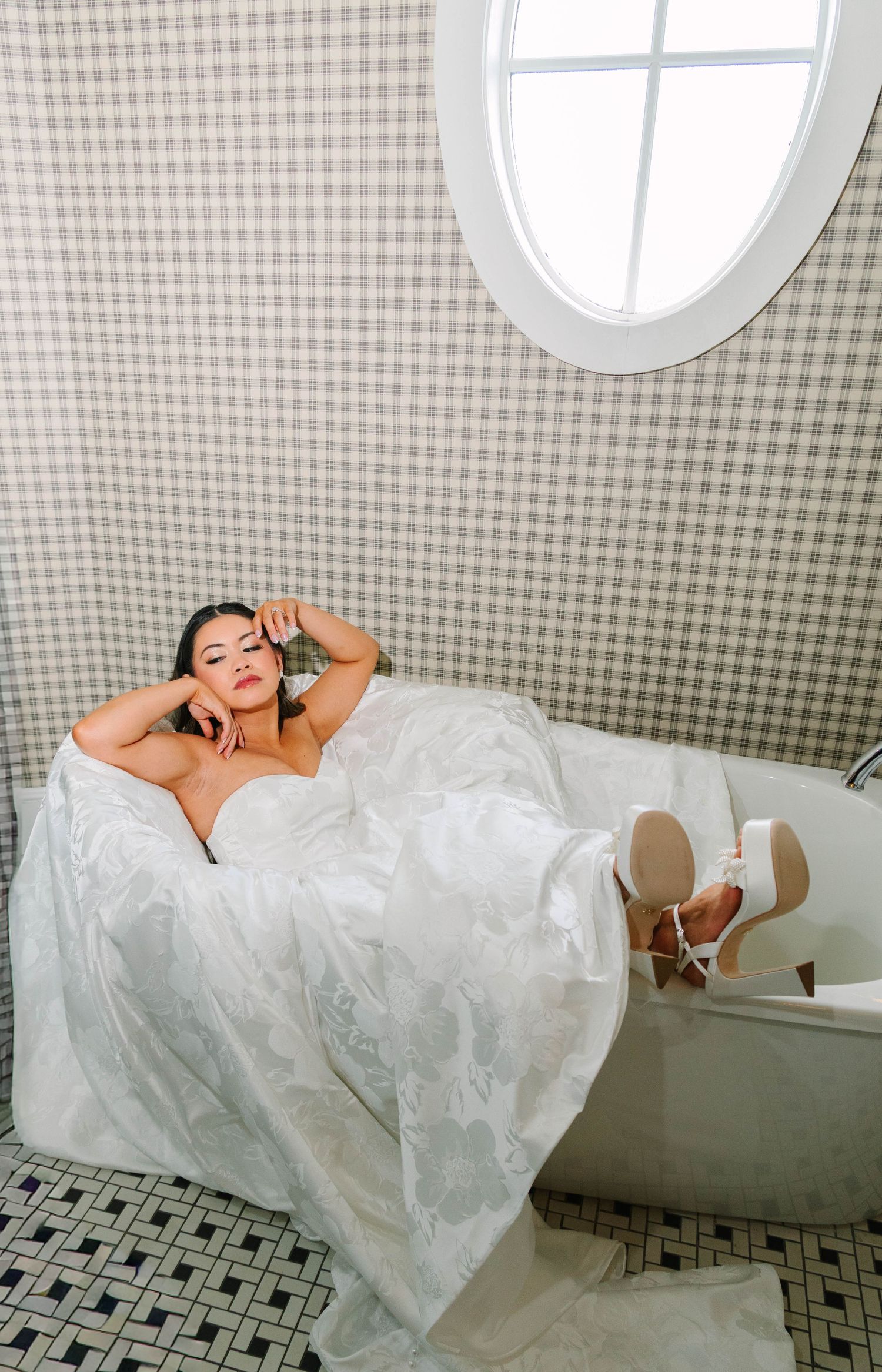 Bride in a white ballgown and heels lounges in a clawfoot bathtub beneath an oval window for bridal portraits.