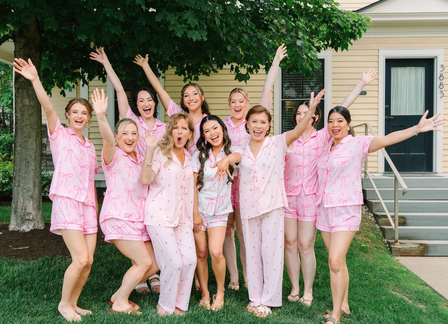 Bride and bridesmaids cheering in matching pink pajamas outside yellow house.