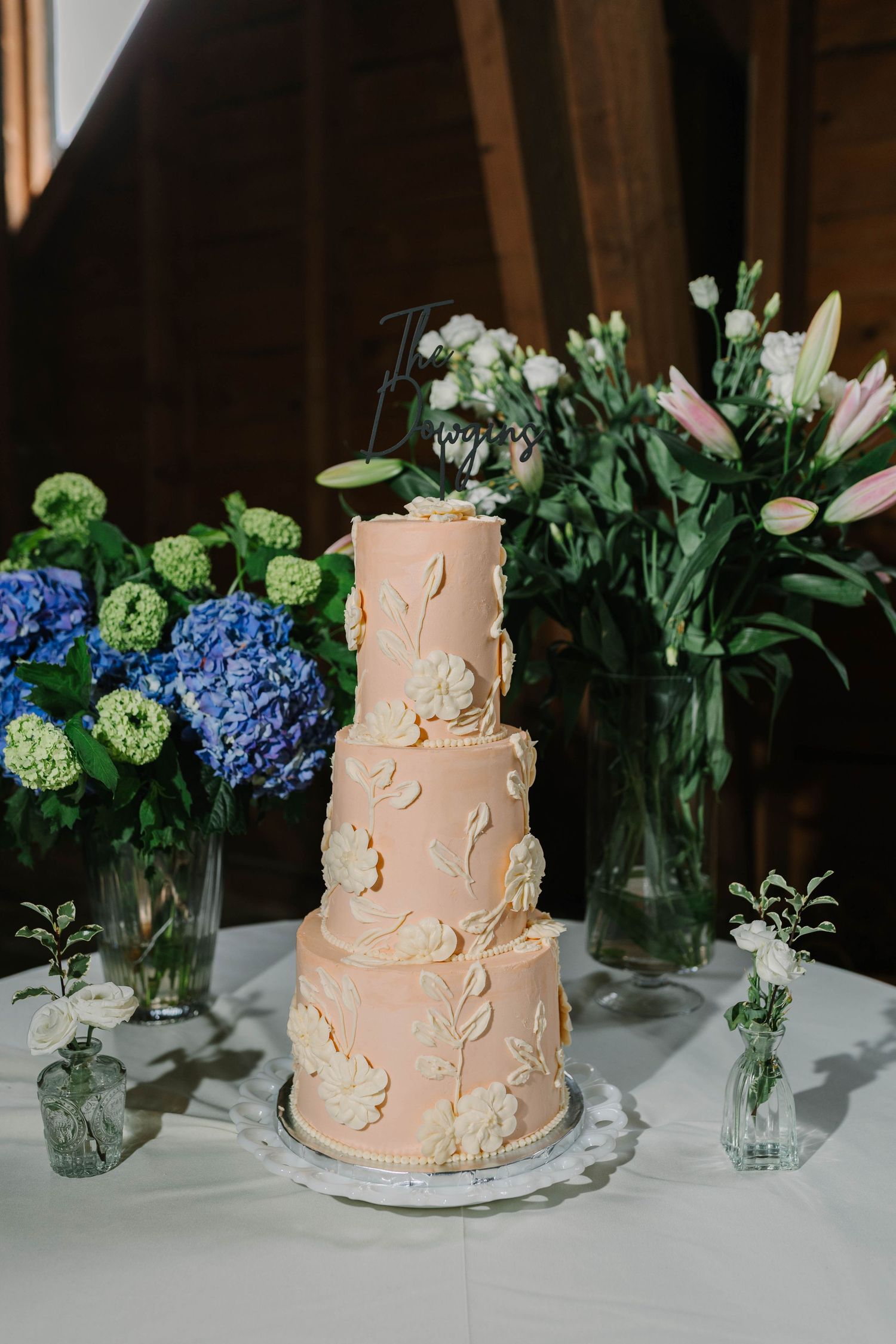 Elegant four-tier blush pink wedding cake with floral details displayed beside blue hydrangeas and lilies.