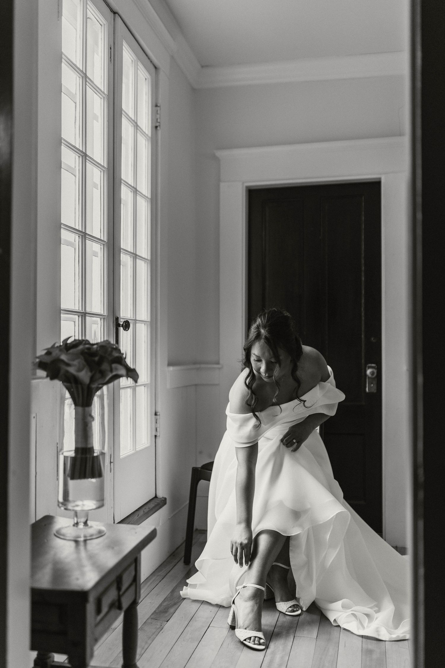 Black and white photo of bride in wedding gown fastening her heels near a bouquet by the door.