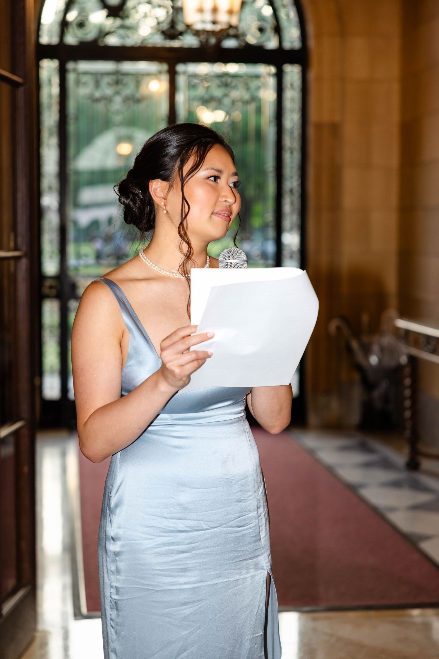 Bridesmaid in silver satin gown delivering speech while holding papers at wedding reception.