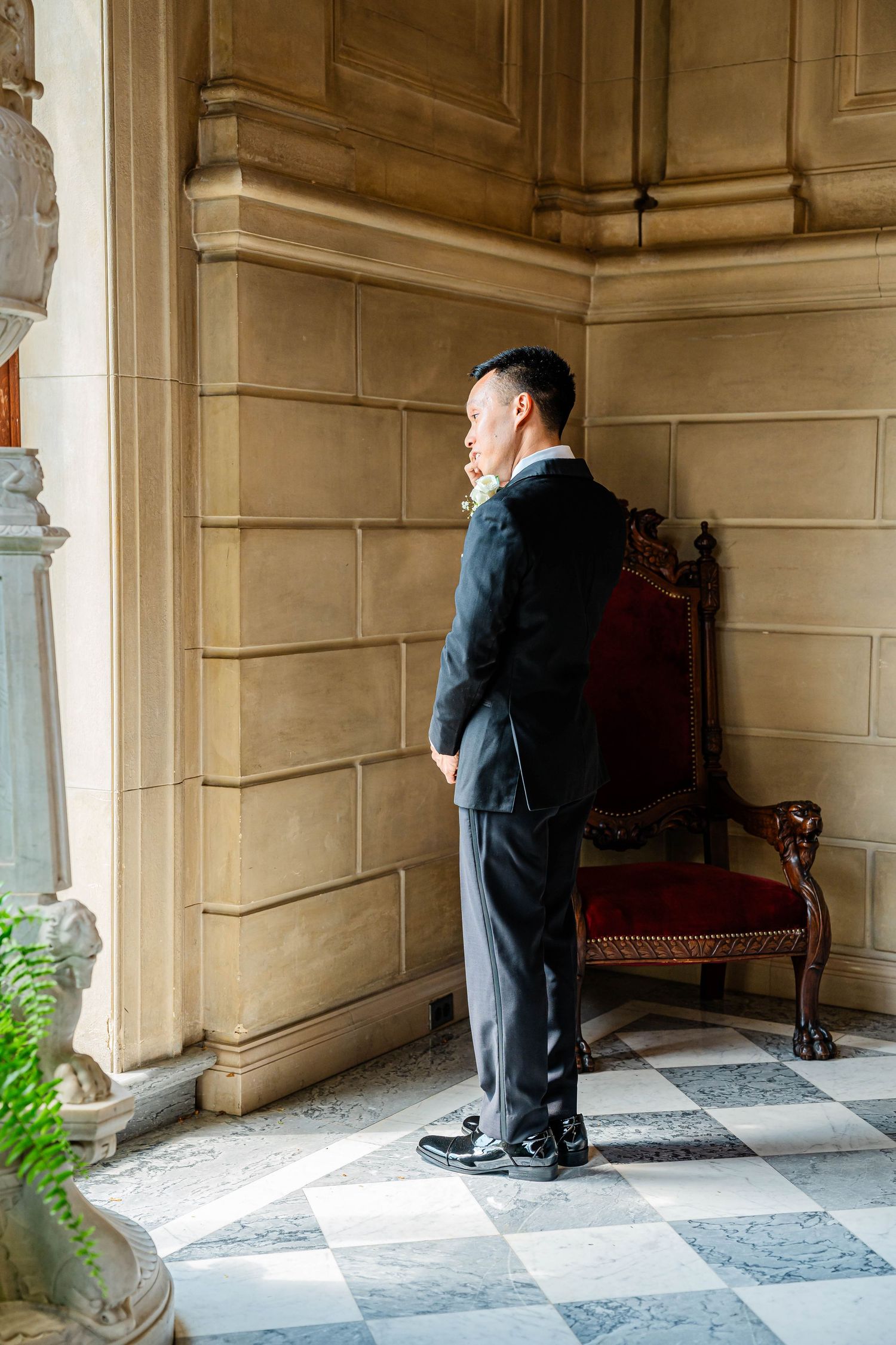 Dapper Asian groom in black suit stands contemplatively beside ornate chair in elegant venue.