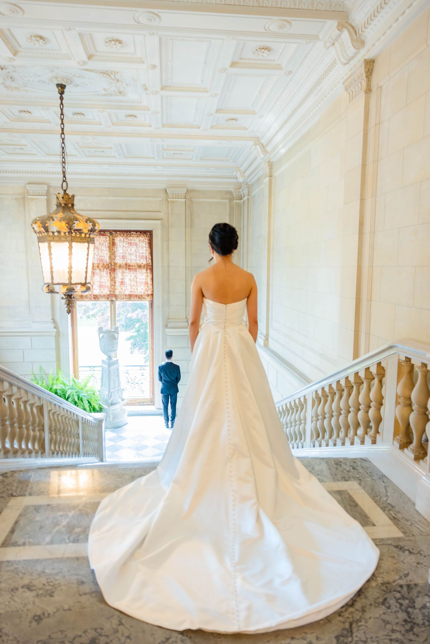 Bride in stunning white cathedral train gown stands in grand marble staircase hall.