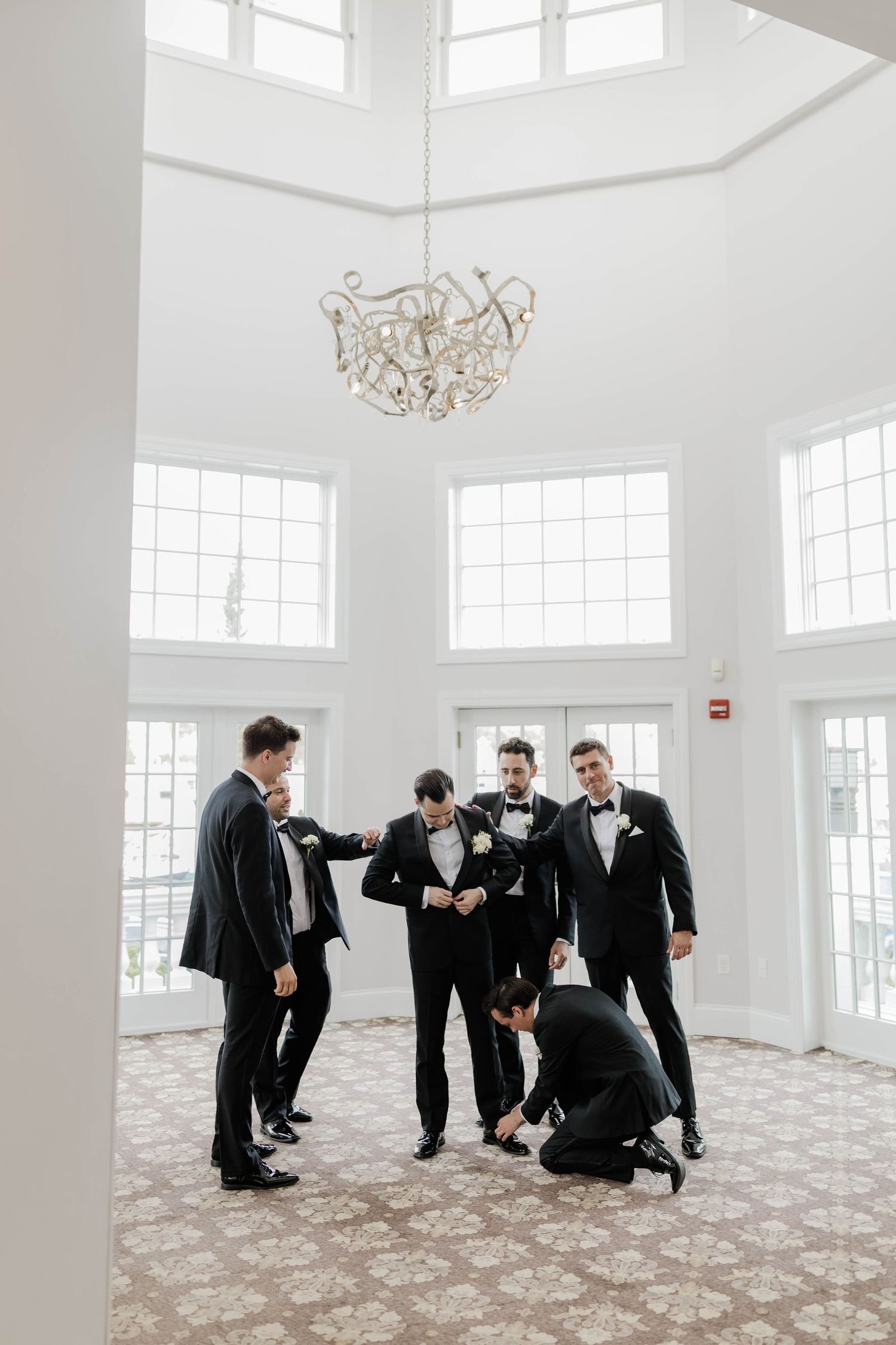 Groomsmen in black tuxedos help each other prepare under chandelier in bright white venue.