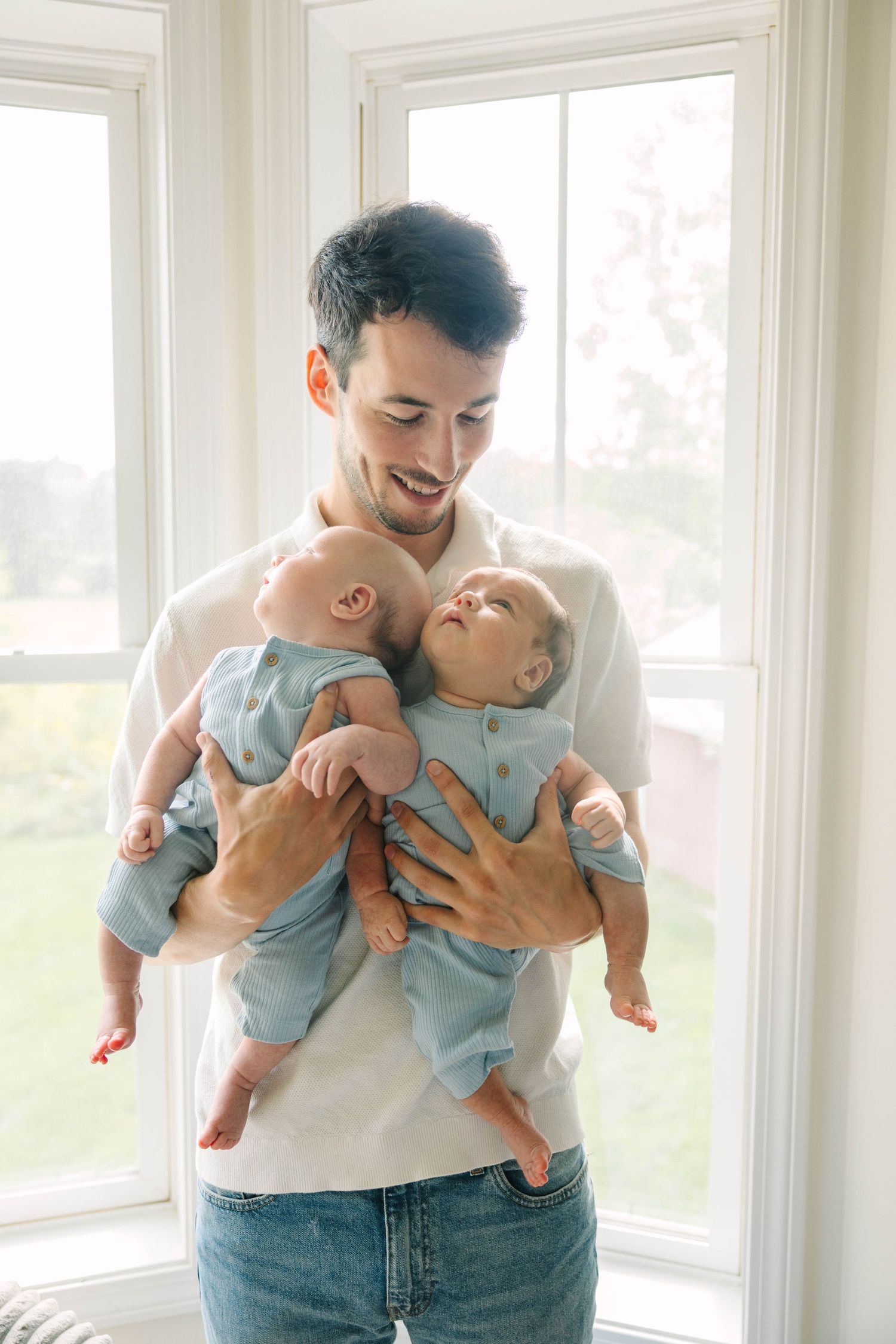 Smiling father holding twin babies in blue outfits near bright sunlit windows.