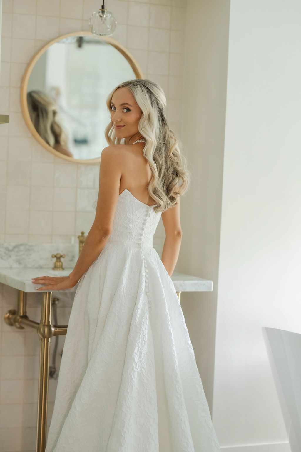 Bride in strapless white lace A-line wedding gown poses in elegant marble bathroom with round gold mirror.