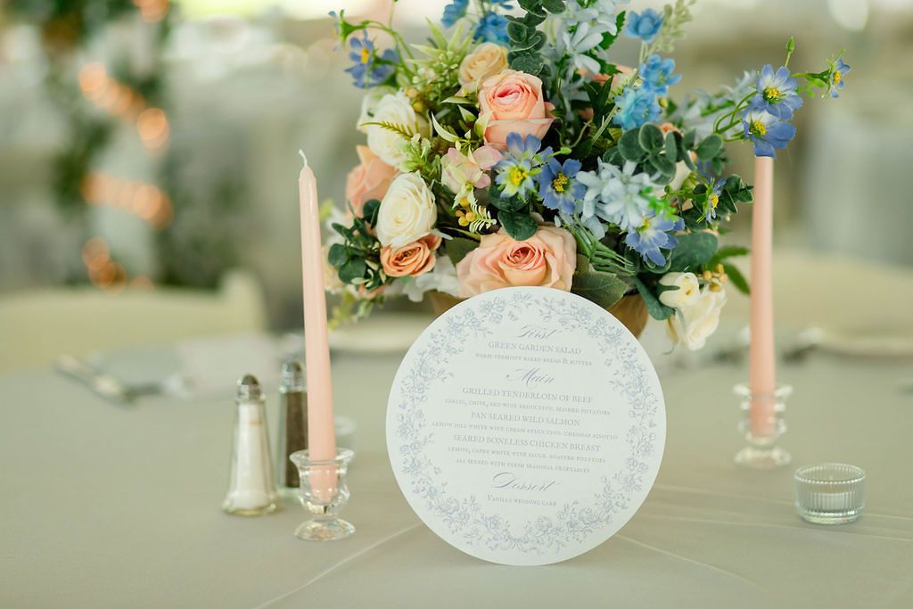 Wedding reception table with peach and blue floral centerpiece, pink taper candles, and round menu card.