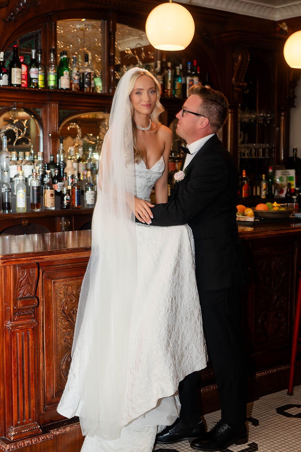 Bride in strapless white gown and veil stands at an ornate wooden bar with the groom in a tuxedo.