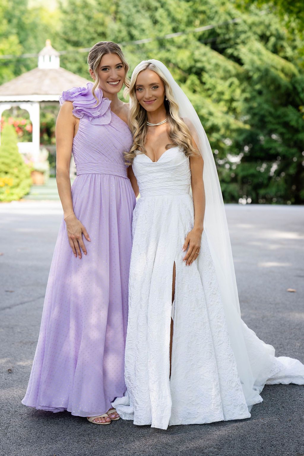Bride in white lace gown poses with bridesmaid in lavender dress outdoors near a gazebo.