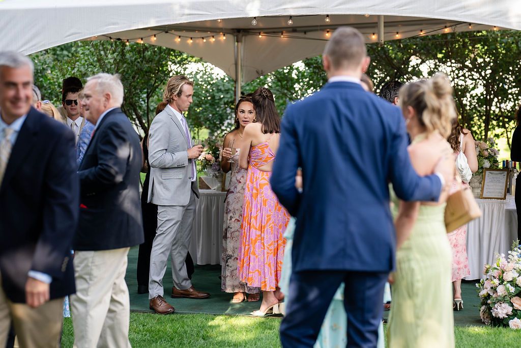 Wedding guests mingling under a white tent with string lights at an outdoor summer reception.