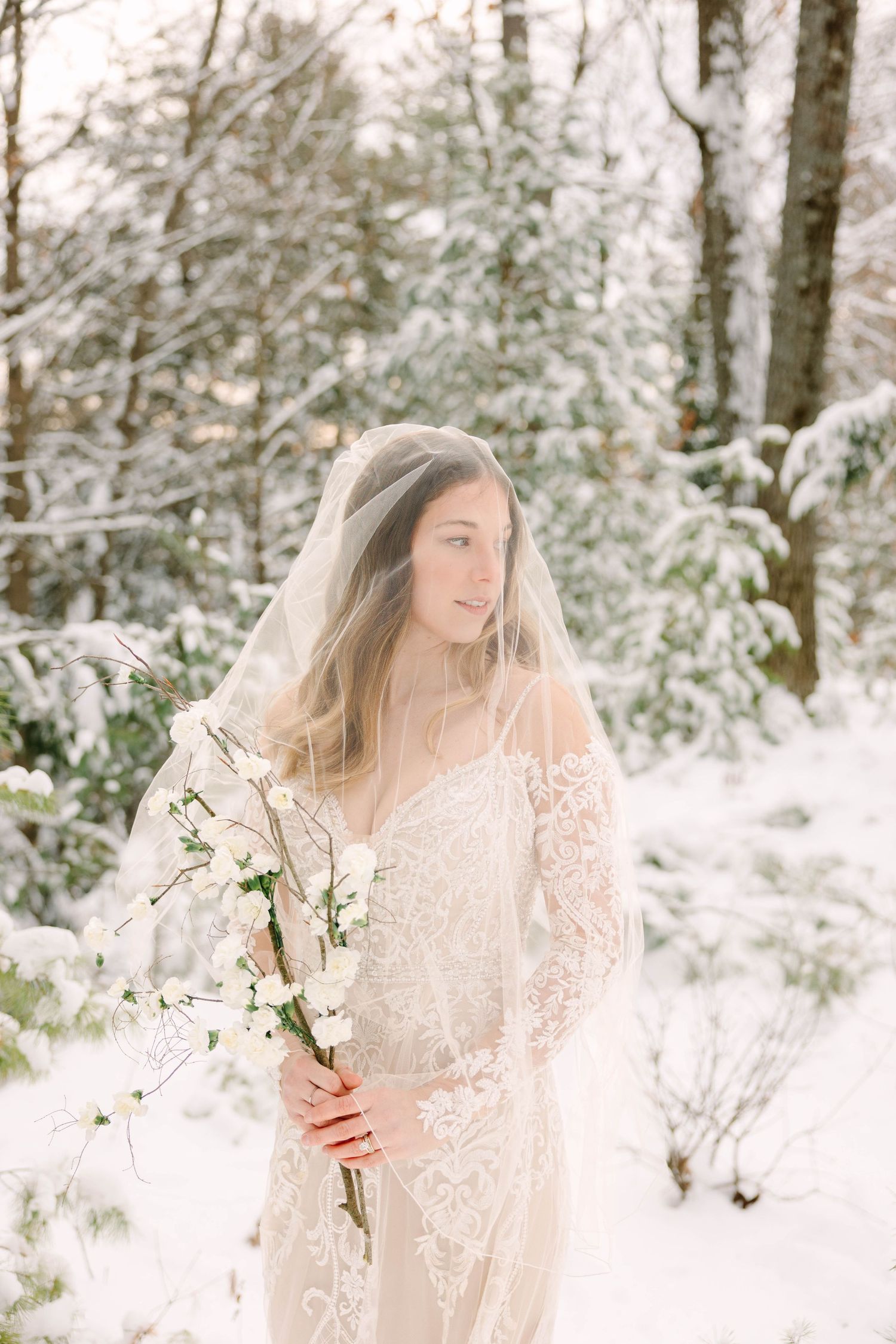 Serene bride in lace gown and sheer veil holding white blooms among snow-covered trees.